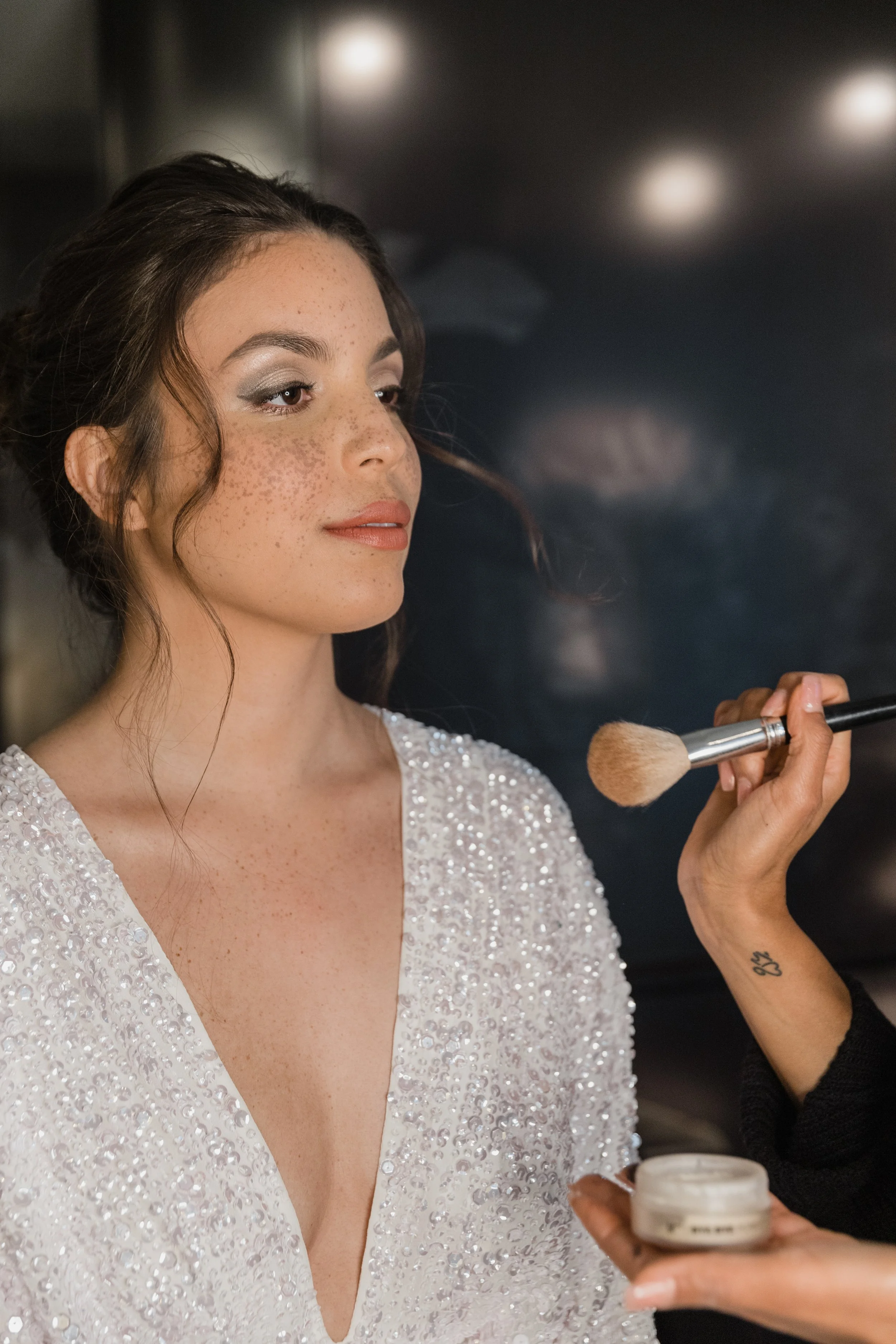 Junge Frau mit Sommersprossen trägt ein silbernes Kleid, während Make-up-Artistinnen sie mit Pinsel schminken.