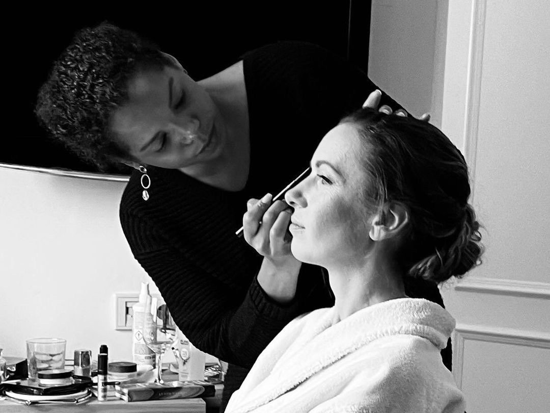 Make-up Artist, die einer Frau beim Schminken auf einem Stuhl hilft, im Raum mit Make-up-Produkten.