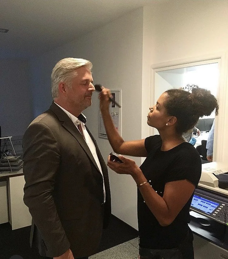 Eine Frau gibt einem Mann im Anzug ein Make-up auf. Sie steht in einem Büro oder einer Arbeitsumgebung.