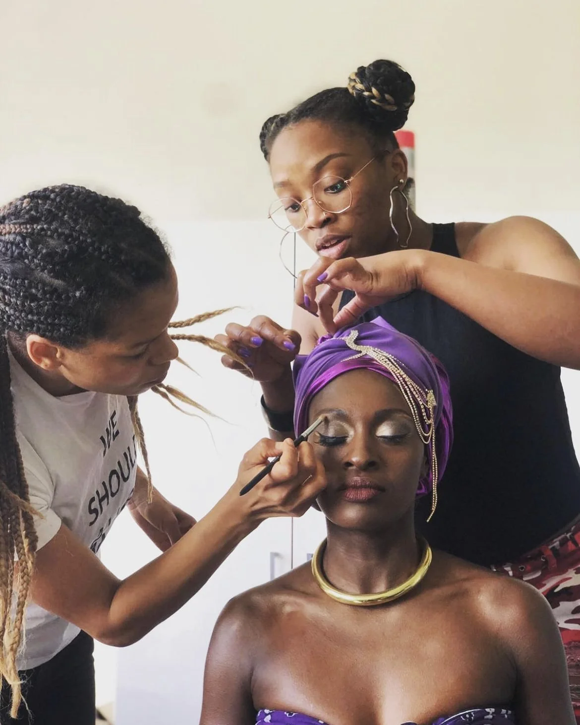 Dreifache Szene: Zwei Frauen arbeiten im Hintergrund an der Makeup- und Haarstyling eines Models im Vordergrund, das einen prachtvollen violetten Kopfschmuck und Schmuck trägt. Die Frau beim Makeup trägt einen weißen T-Shirt, die andere trägt Brillen und große Ohrringe.