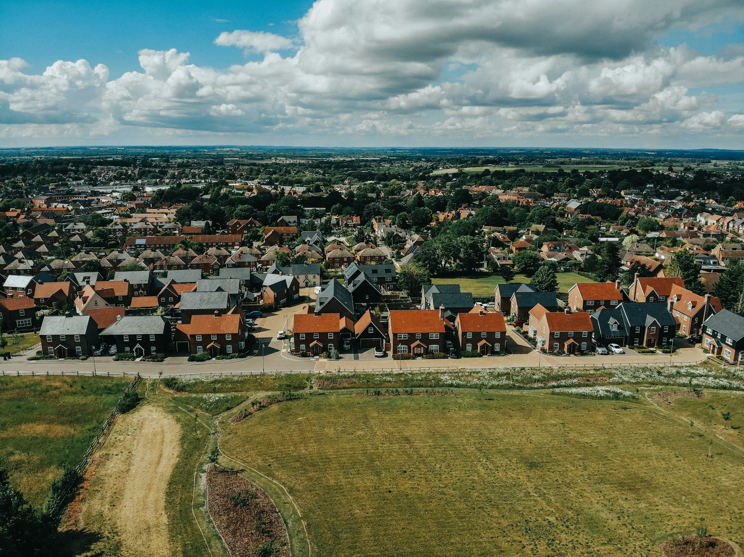 Aerial view of a suburban neighborhood with houses, green trees, and wide-open fields under a partly cloudy sky.