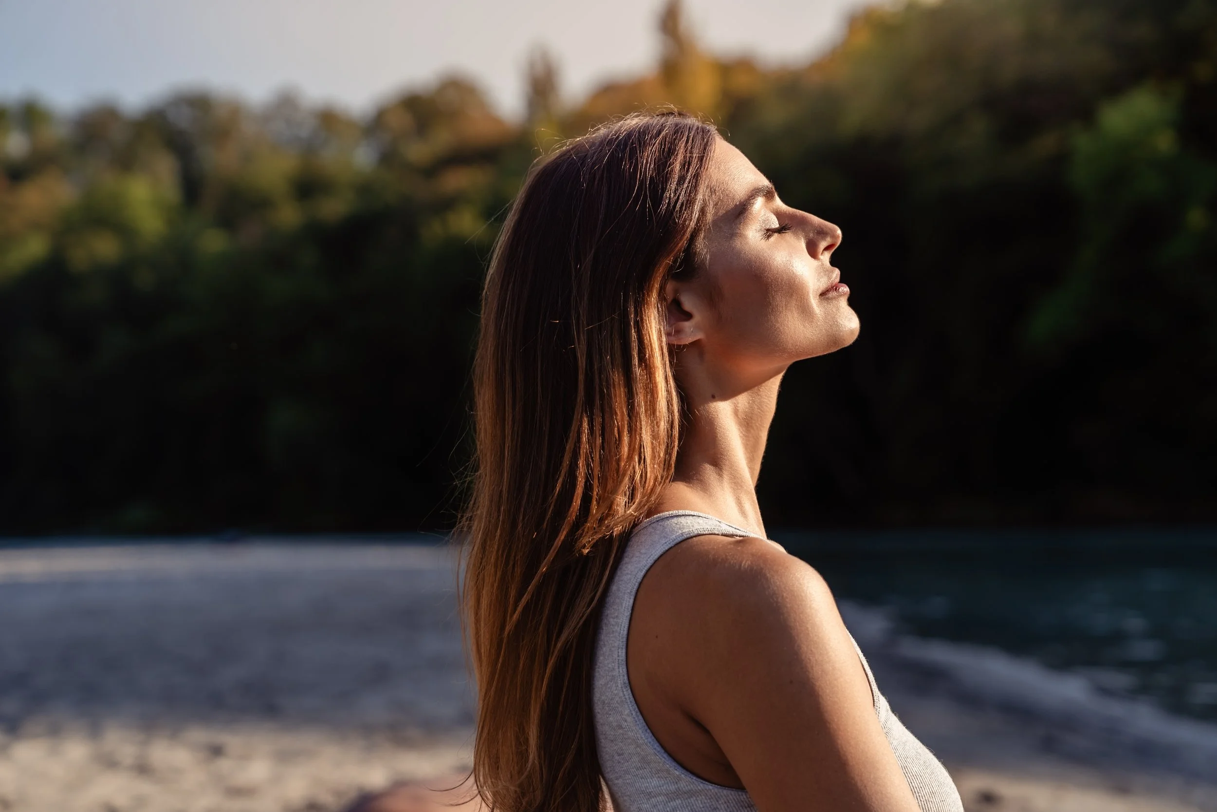 Woman with long brown hair meditating outdoors with eyes closed, facing the sun.