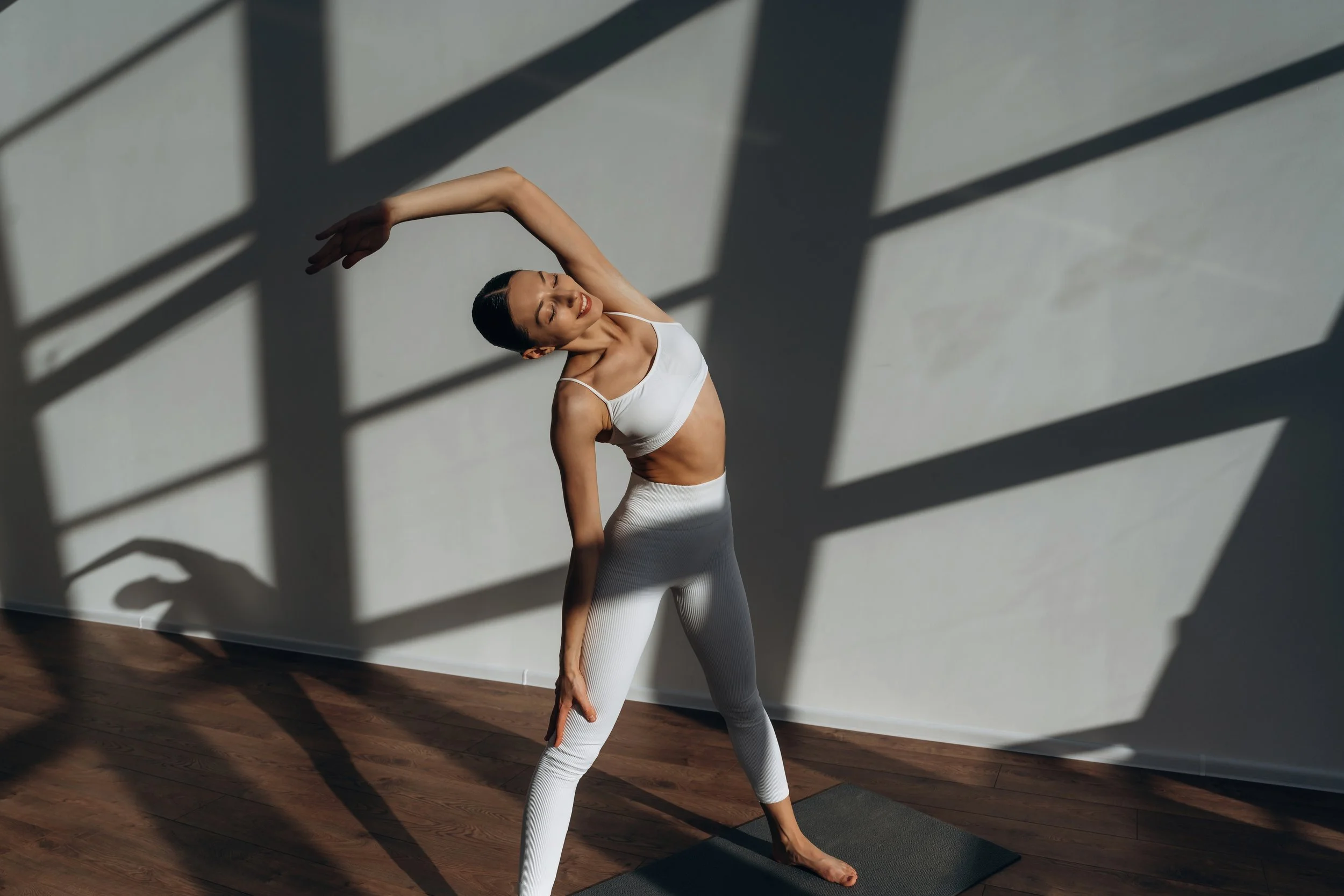 A woman practicing yoga indoors, standing in a side stretching pose with her eyes closed, wearing white athletic wear, and casting shadows on the wall behind her.