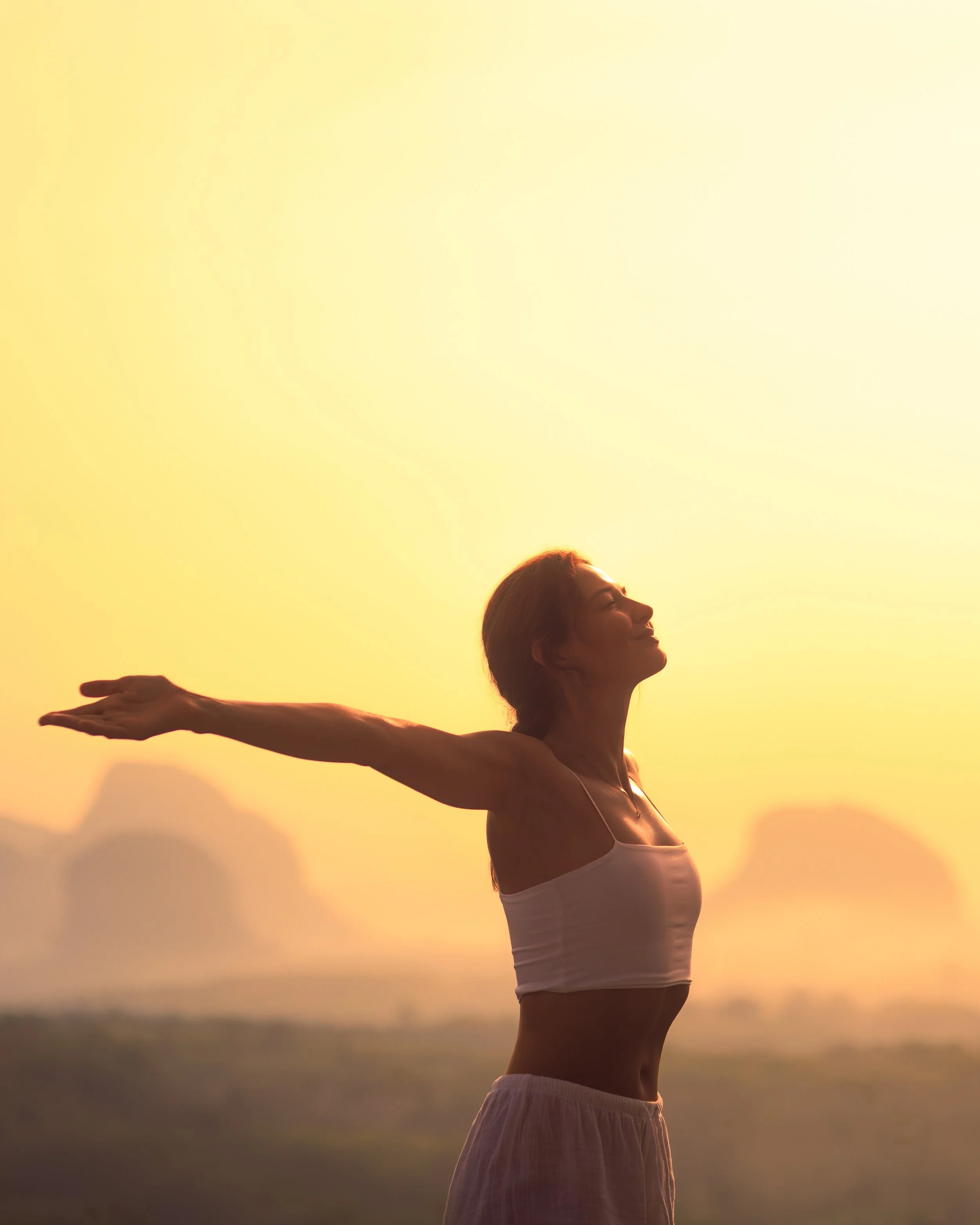 A woman standing outdoors during sunset, with her arms stretched out to the sides, smiling and looking upwards.