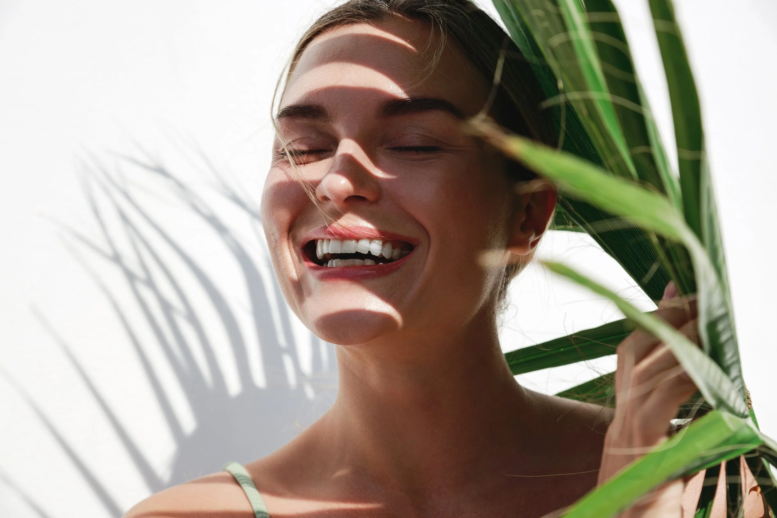 A woman smiling with closed eyes, holding green palm leaves near her face, sunlight casting shadows on her face and the white background.