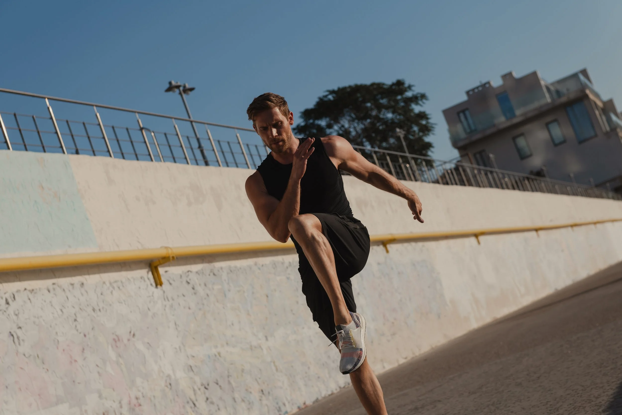 A man in black athletic clothing and sneakers running on an outdoor track under clear blue sky.