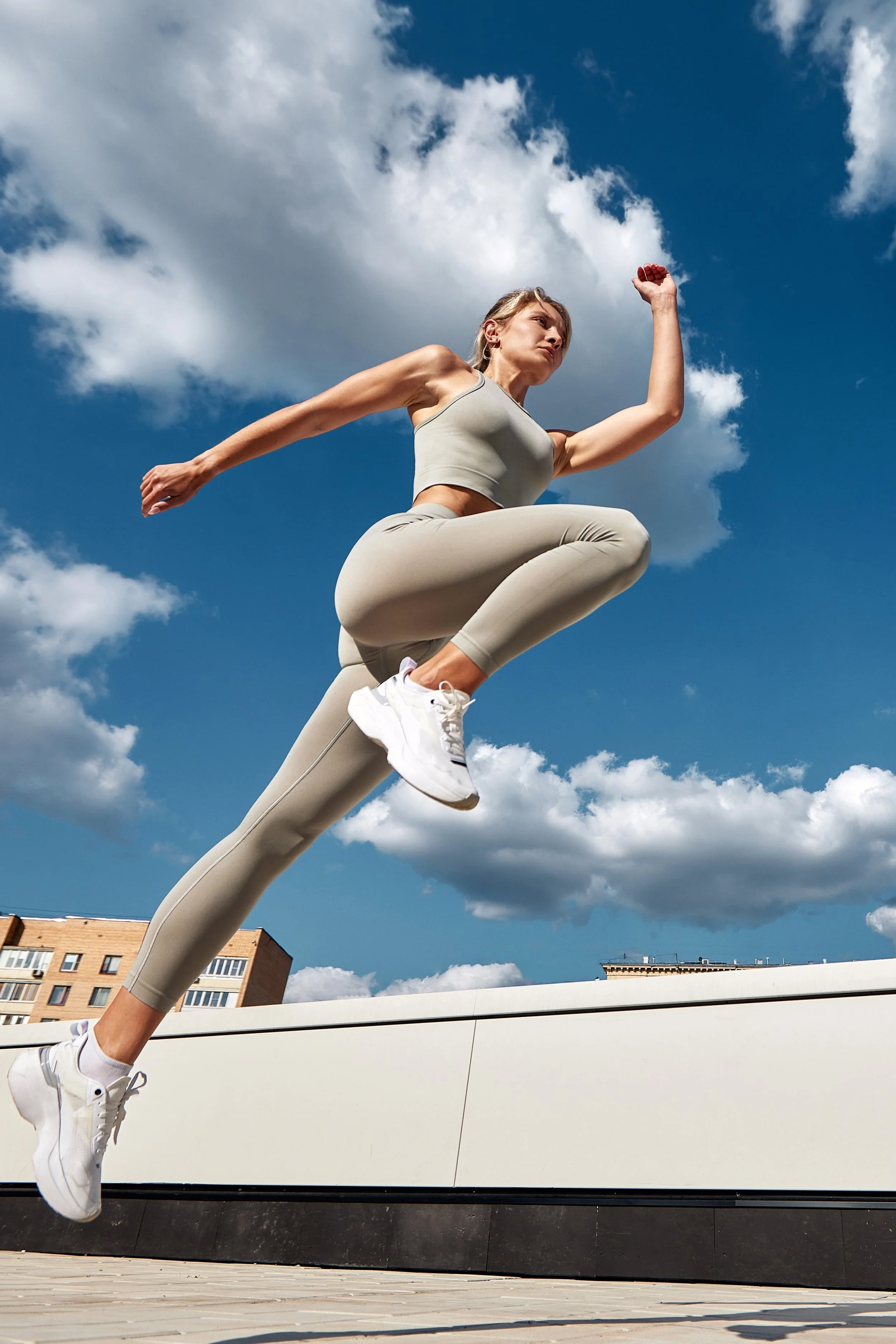 A woman in athletic clothing is jumping in the air outdoors on a rooftop with a cityscape and cloudy sky in the background.