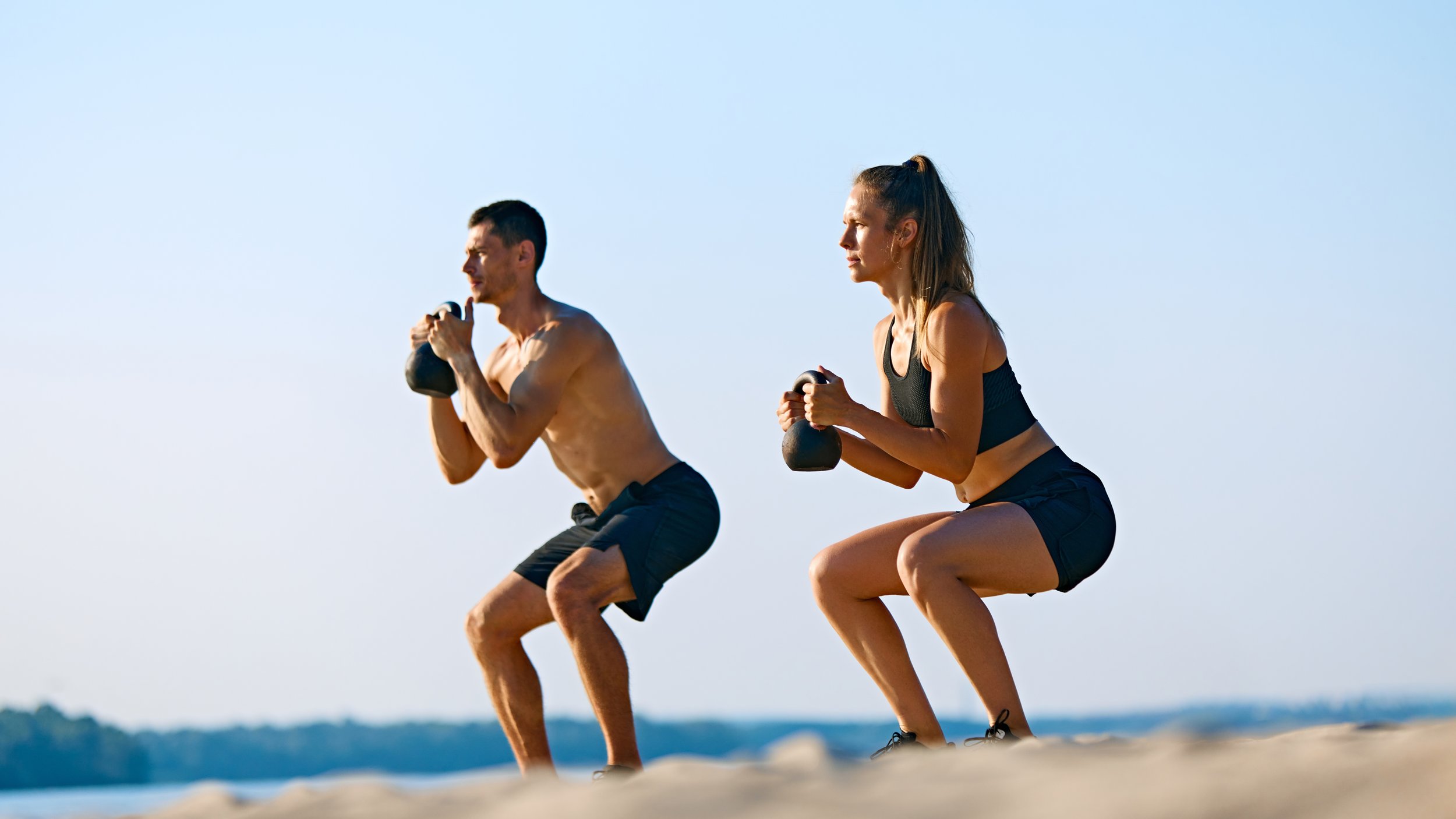 A man and woman exercising outdoors on the beach, performing squats with kettlebells during daytime.