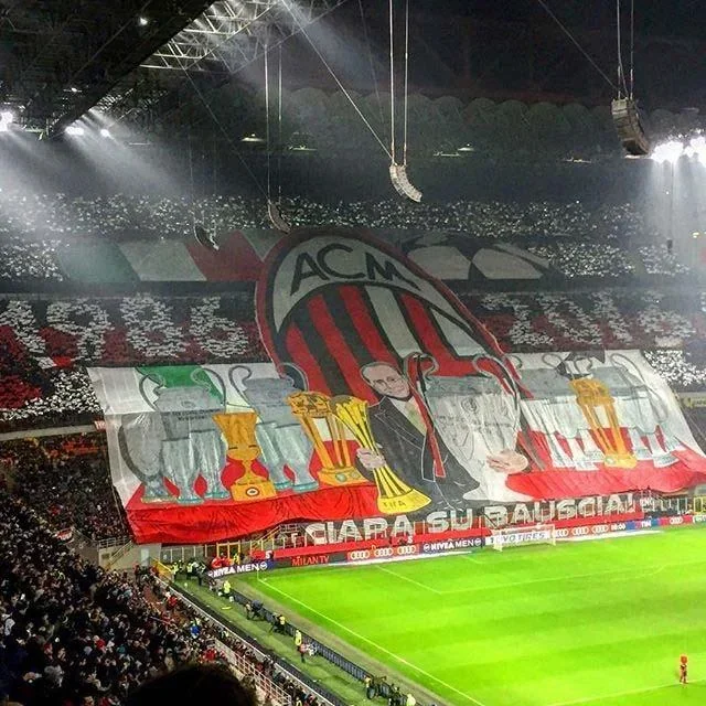 The San Siro stadium filled with AC Milan fans, featuring a massive banner displaying a cartoon character holding multiple trophies, with the AC Milan logo and colourful artwork.