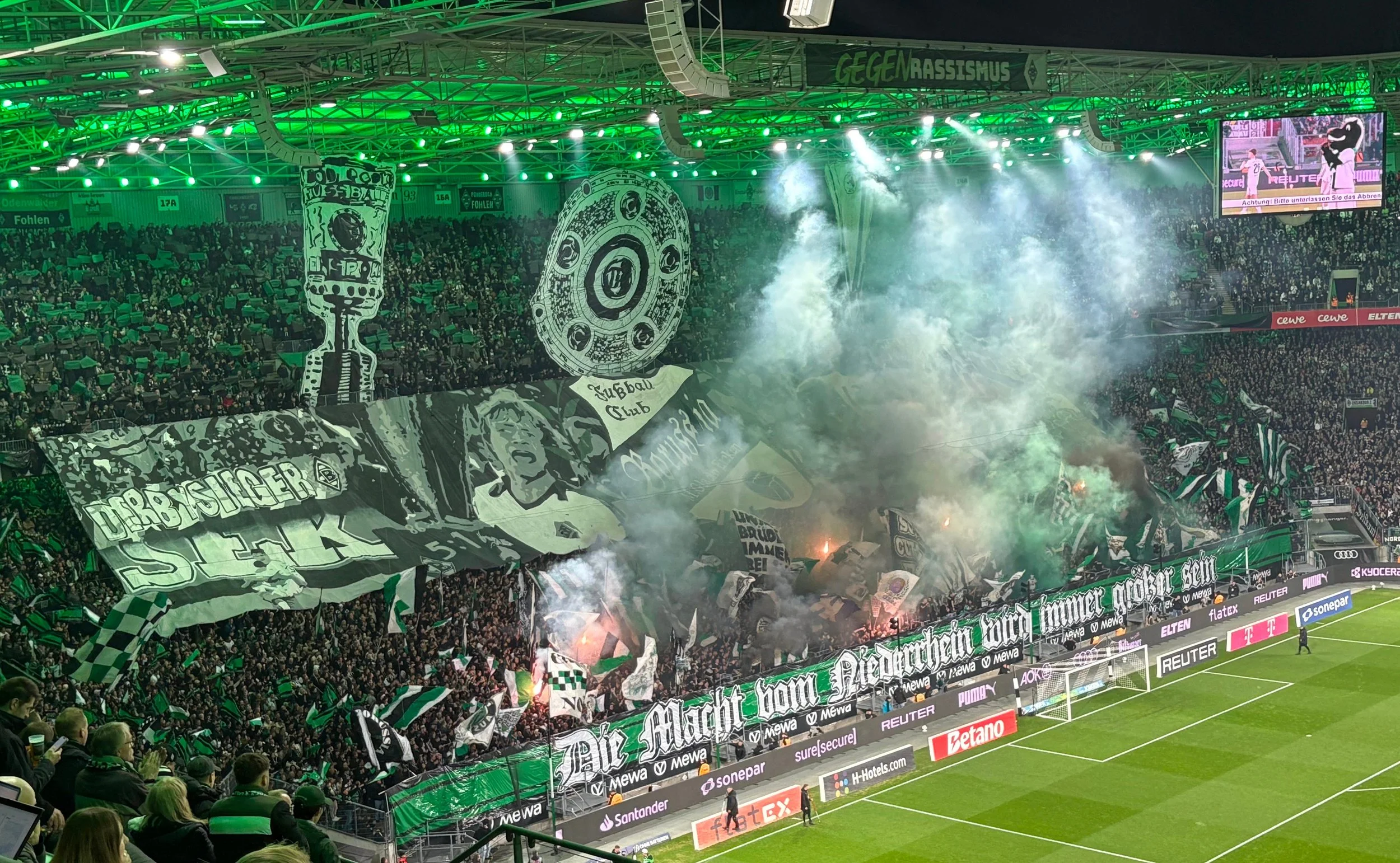 A crowded football stadium filled with Wolfsburg fans in Germany displaying green and black banners and flags. Smoke and bright stadium lights create an energetic atmosphere. Digital screens show a football match.