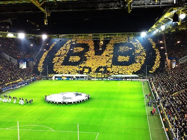 A packed football stadium with fans holding up yellow and black cards to spell out 'BVB' and a large Borussia Dortmund emblem on the seating area, preparing for a match or event. Signal Iduna Park