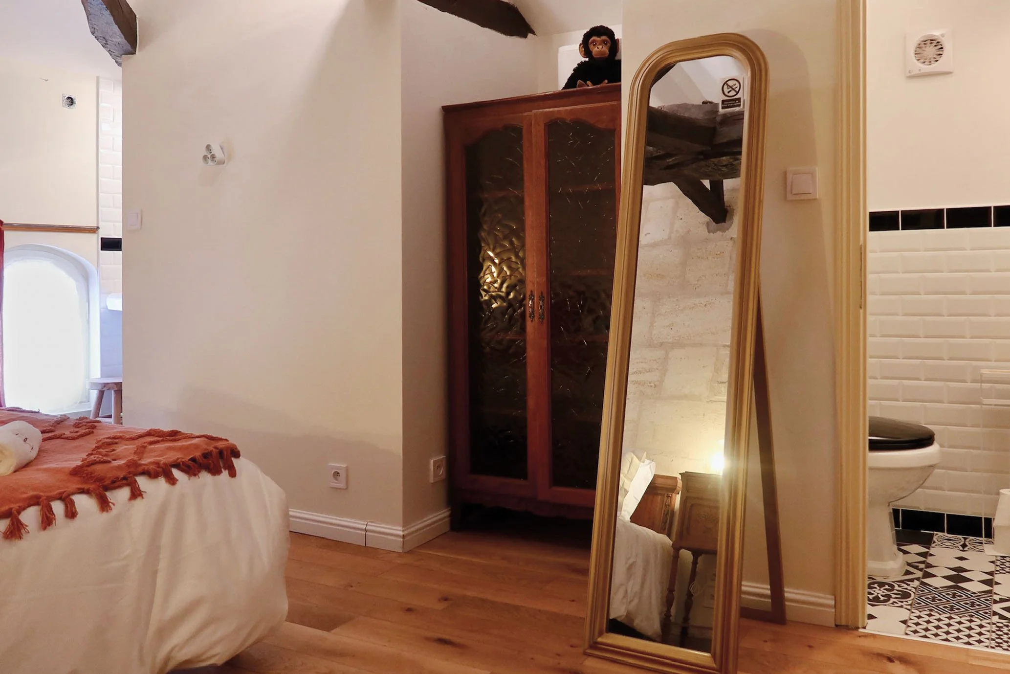 Une pièce de chambre avec un lit à gauche, une grande armoire en bois au fond et un miroir debout à droite, avec un WC visible dans une pièce attenante.