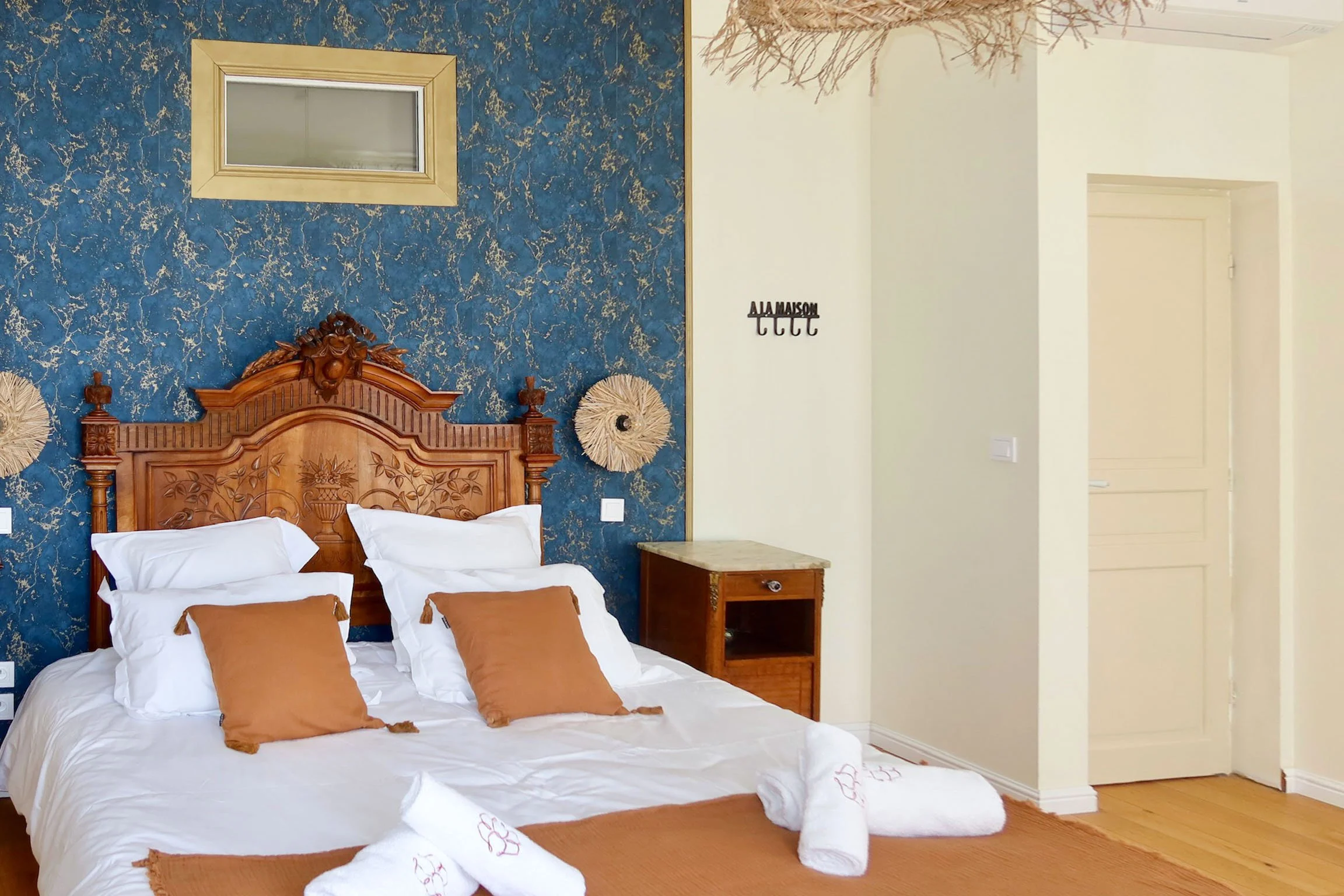 Chambre avec lit en bois sculpté, draps blancs et oreillers marron, mur bleu avec effets marbrés, décoration en paille et cadre au mur, côté droit une porte et un mur blanc avec un crochet mural.