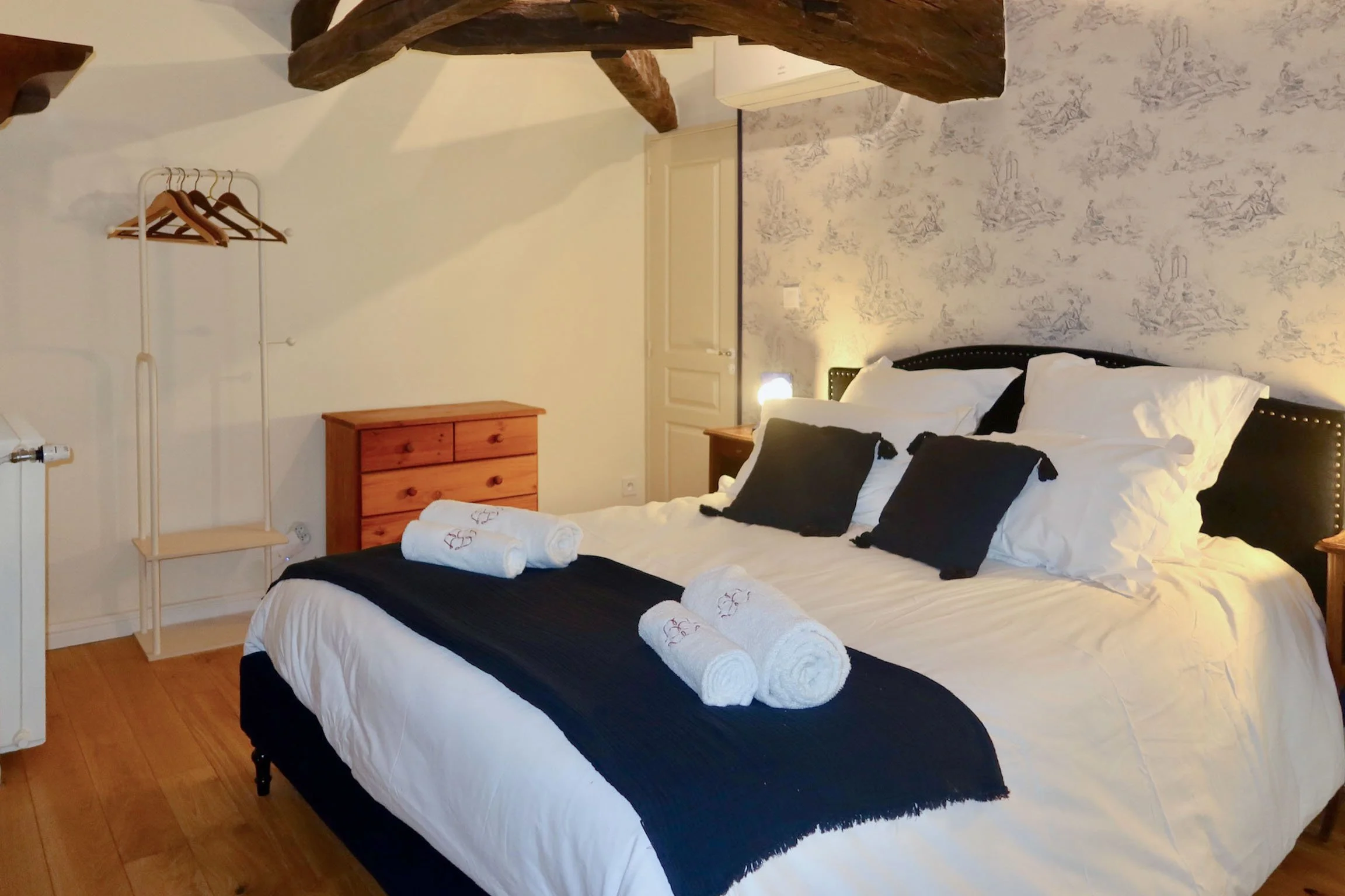 Une chambre avec un lit fait, des serviettes blanches roulées, un z-clou en tissu noir, un meuble en bois, une tête de lit noire avec des clous décoratifs et un mur avec un papier peint à motifs.