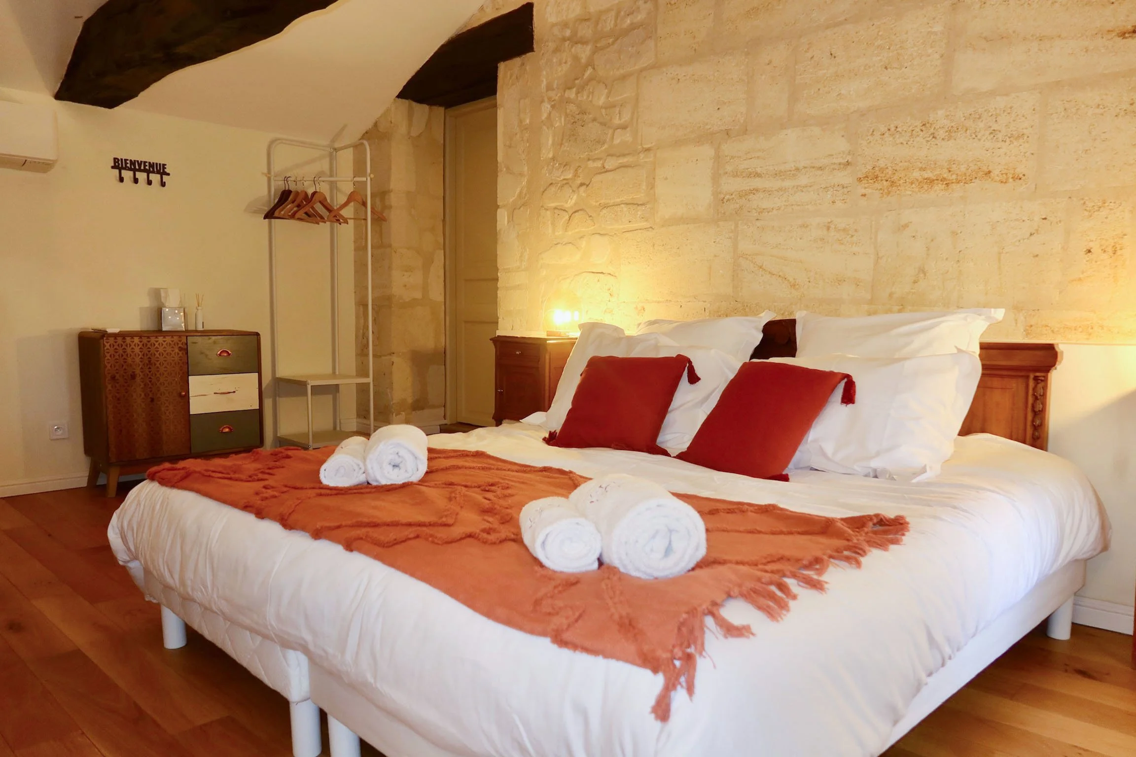 Chambre avec lit double, oreillers blancs et coussins rouges, serviettes blanches roulées sur le lit, mur en pierre beige, petite lampe allumée, meuble en bois, porte en bois, étendoir à vêtements, décoration simple, atmosphère chaleureuse.