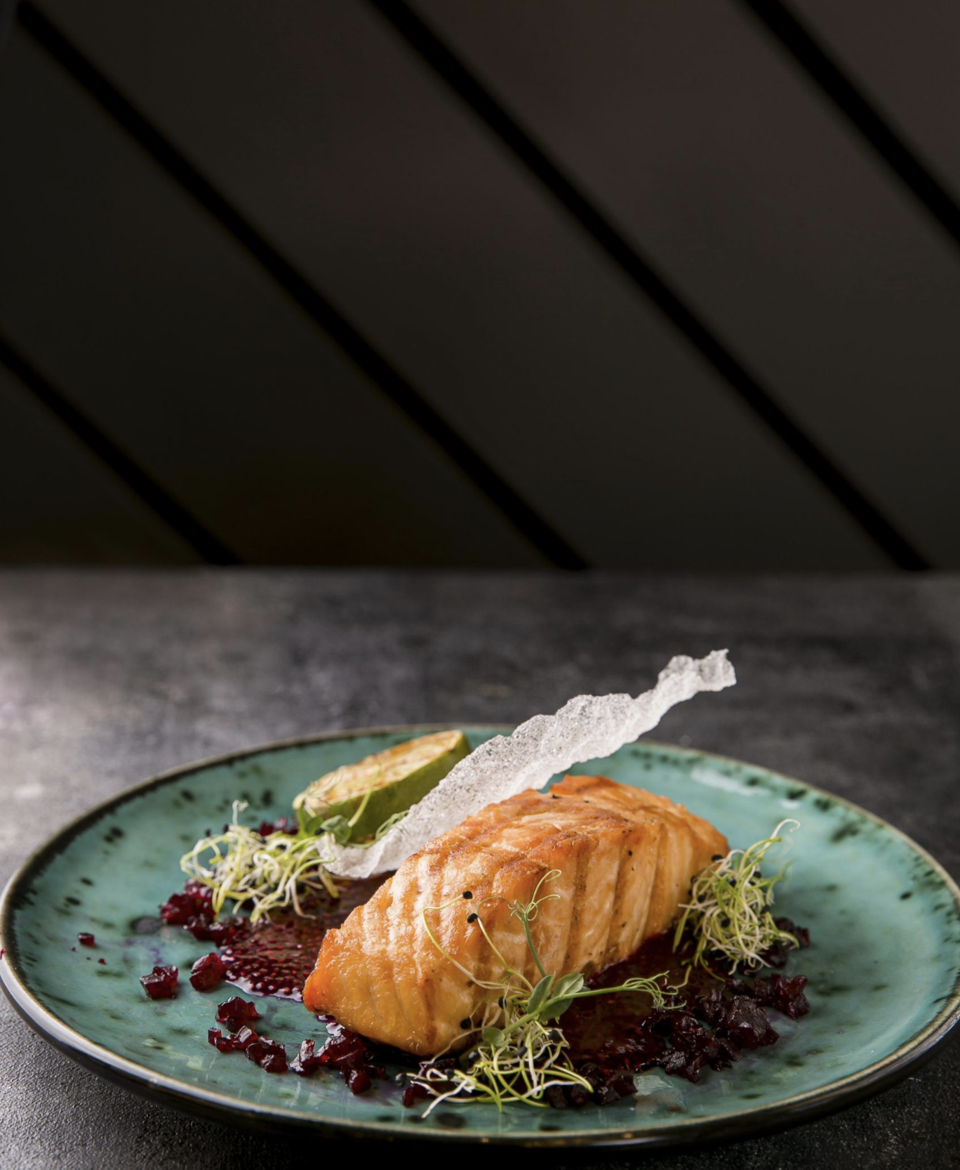 Filet de saumon cuit, garni de micro-verts, accompagné de sauce et décoré de radis noirs, servi sur une assiette en céramique turquoise, avec des éléments croustillants et une tranche de concombre.