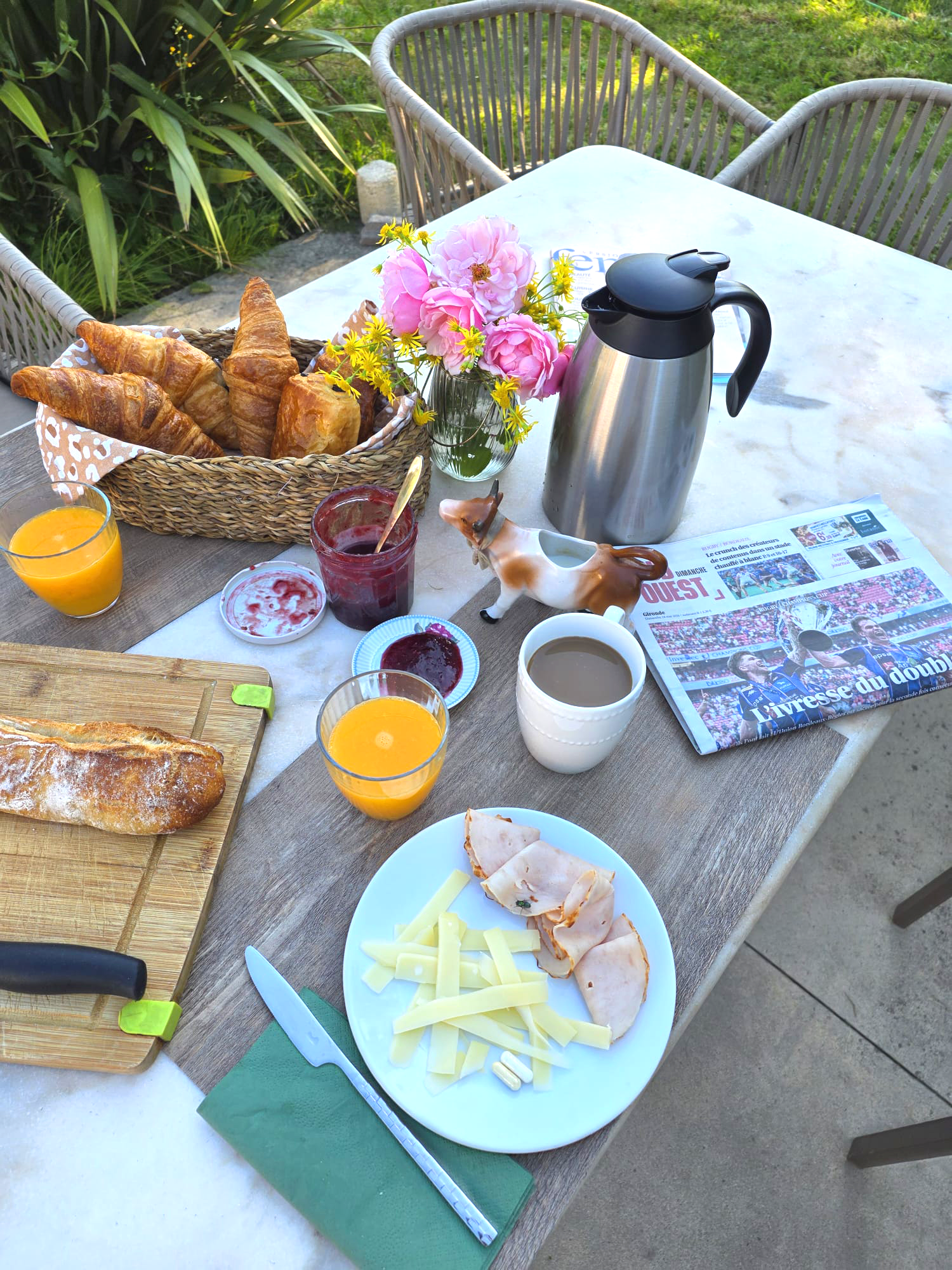 Un petit déjeuner avec croissants, pain, jus d'orange, confiture, fromage, jambon, une cafetière, un vase de fleurs, un journal, et une figure de cheval en céramique sur une table en bois à l'extérieur.