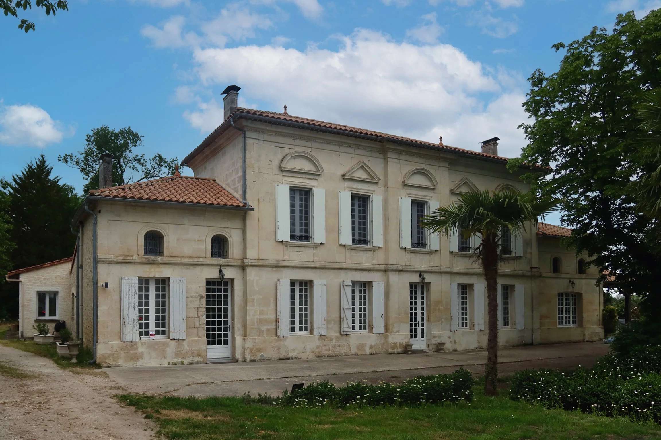 Une grande maison en pierre avec plusieurs fenêtres à volets blancs, un toit en tuiles rouges, entourée d'arbres et d'un jardin avec des fleurs, sous un ciel bleu avec quelques nuages.