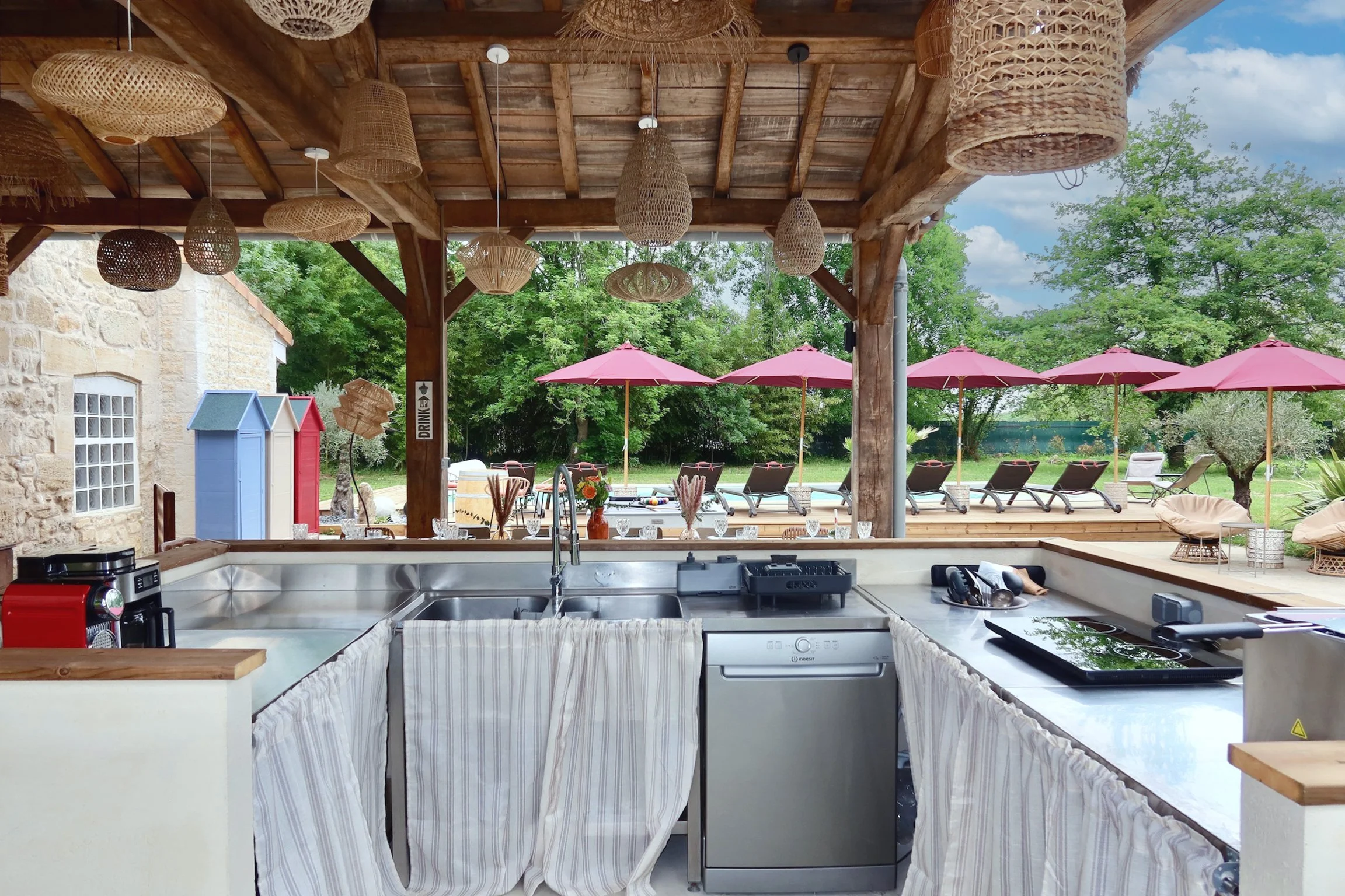 Une cuisine ouverte donnant sur une terrasse extérieure avec des parasols roses et des chaises longues. Le plafond est en bois avec des lampes en rotin suspendues.