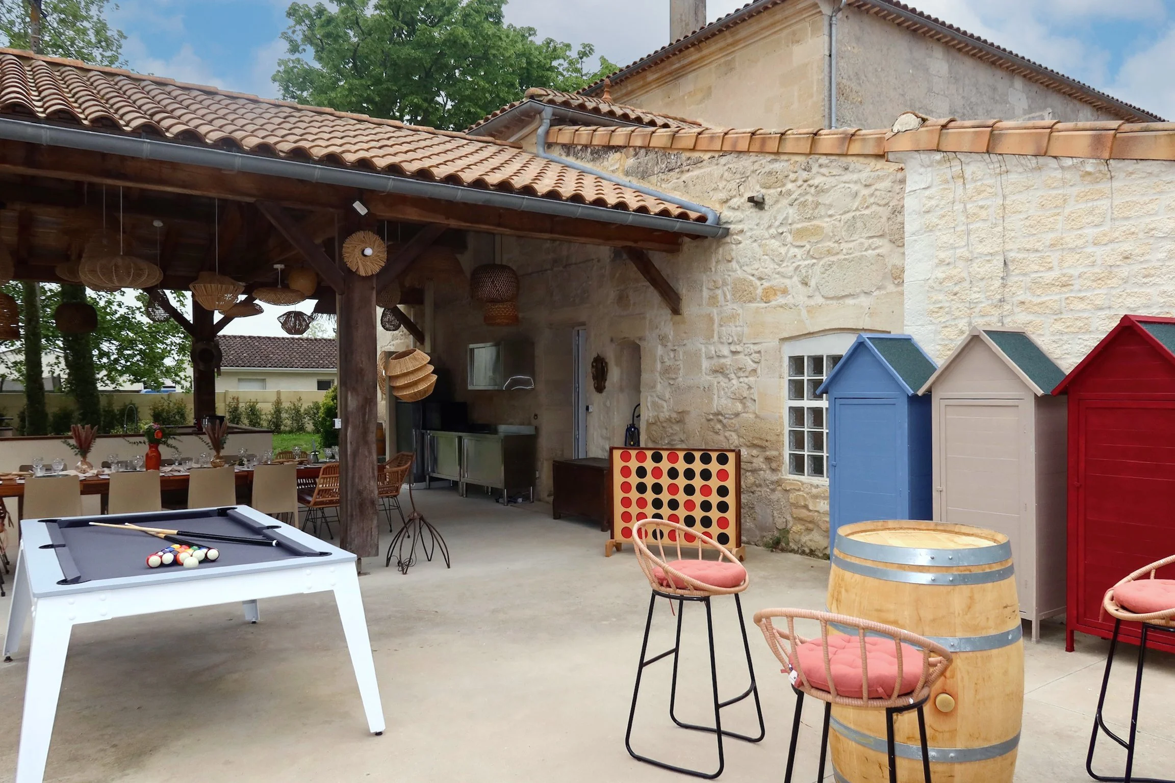 Espace extérieur avec mobilier de style champêtre, une table de billard, chaises en rotin avec coussins, un bar en tonneau, et des jeux de société, sous un toit en bois et tuiles, avec un mur en pierre et des petits maisons en bois colorées.