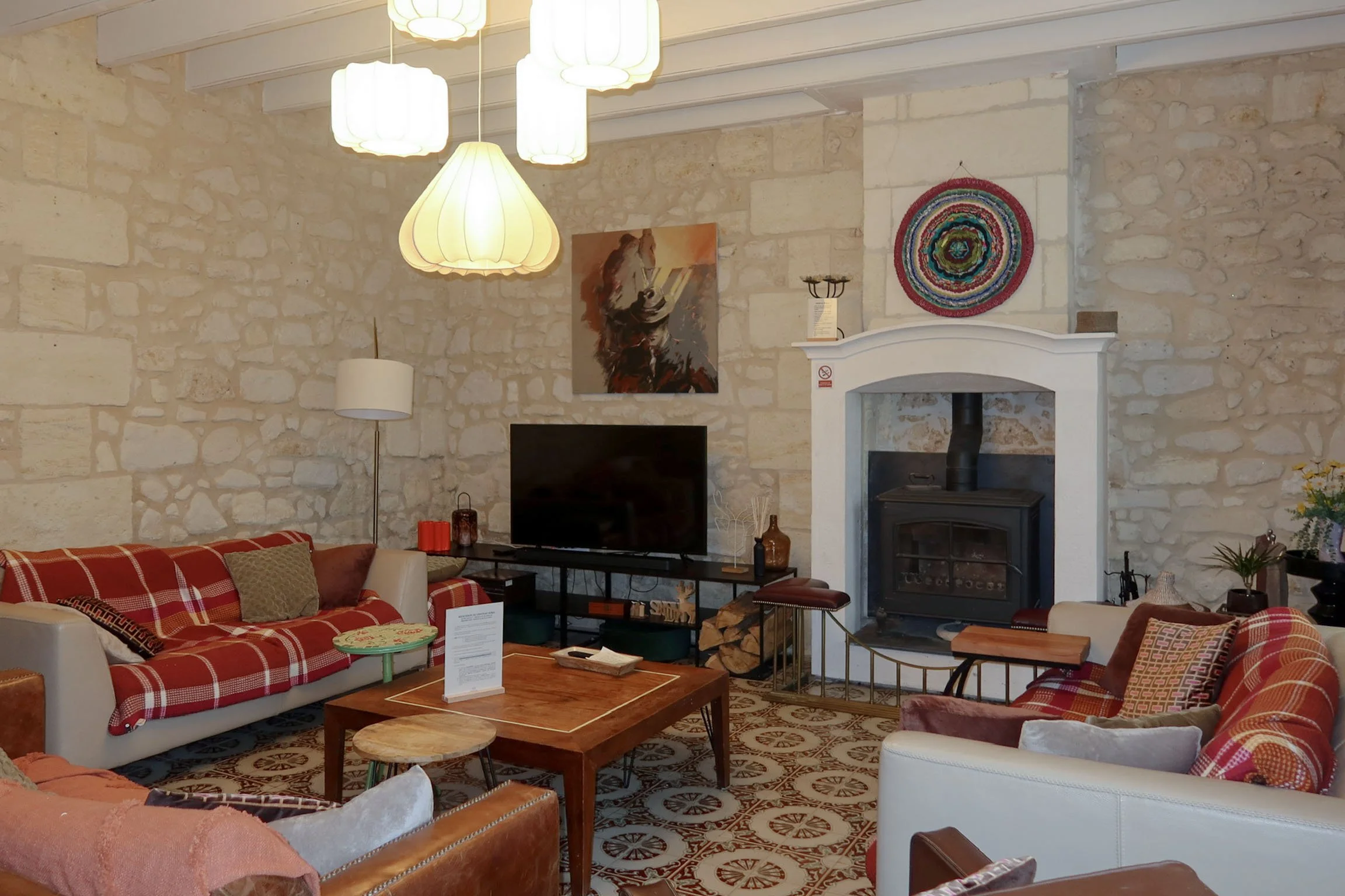 Intérieur d'un salon avec murs en pierre beige, plusieurs lampes suspendues au plafond, un canapé à carreaux rouge et blanc, une cheminée en pierre blanche décorée d'une œuvre d'art, une table basse en bois, et des meubles et objets décoratifs divers.