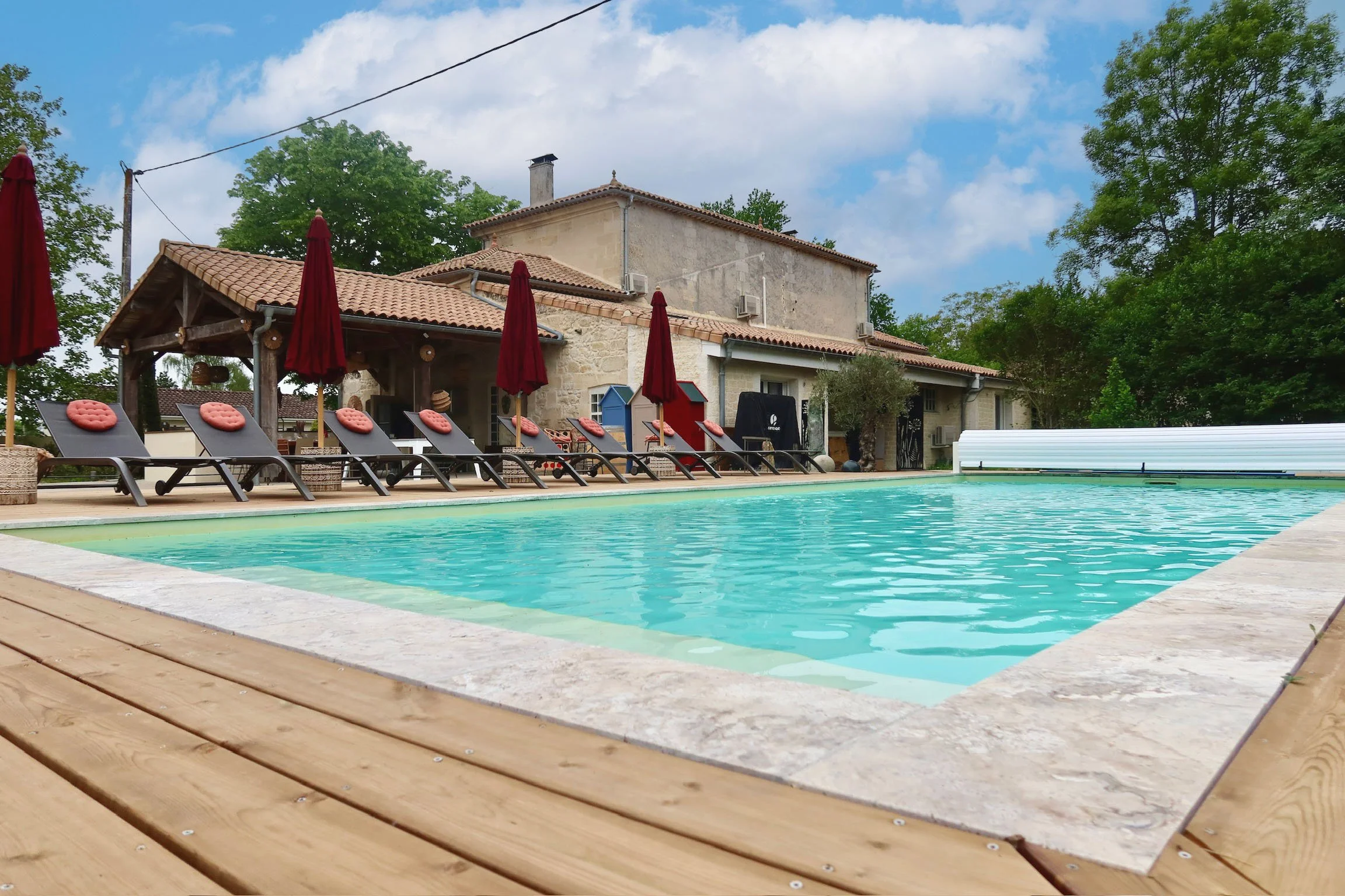 Piscine extérieure avec chaise longues, parasols rouges, maison en pierres avec toit en tuiles, arbres verts et ciel bleu avec quelques nuages.