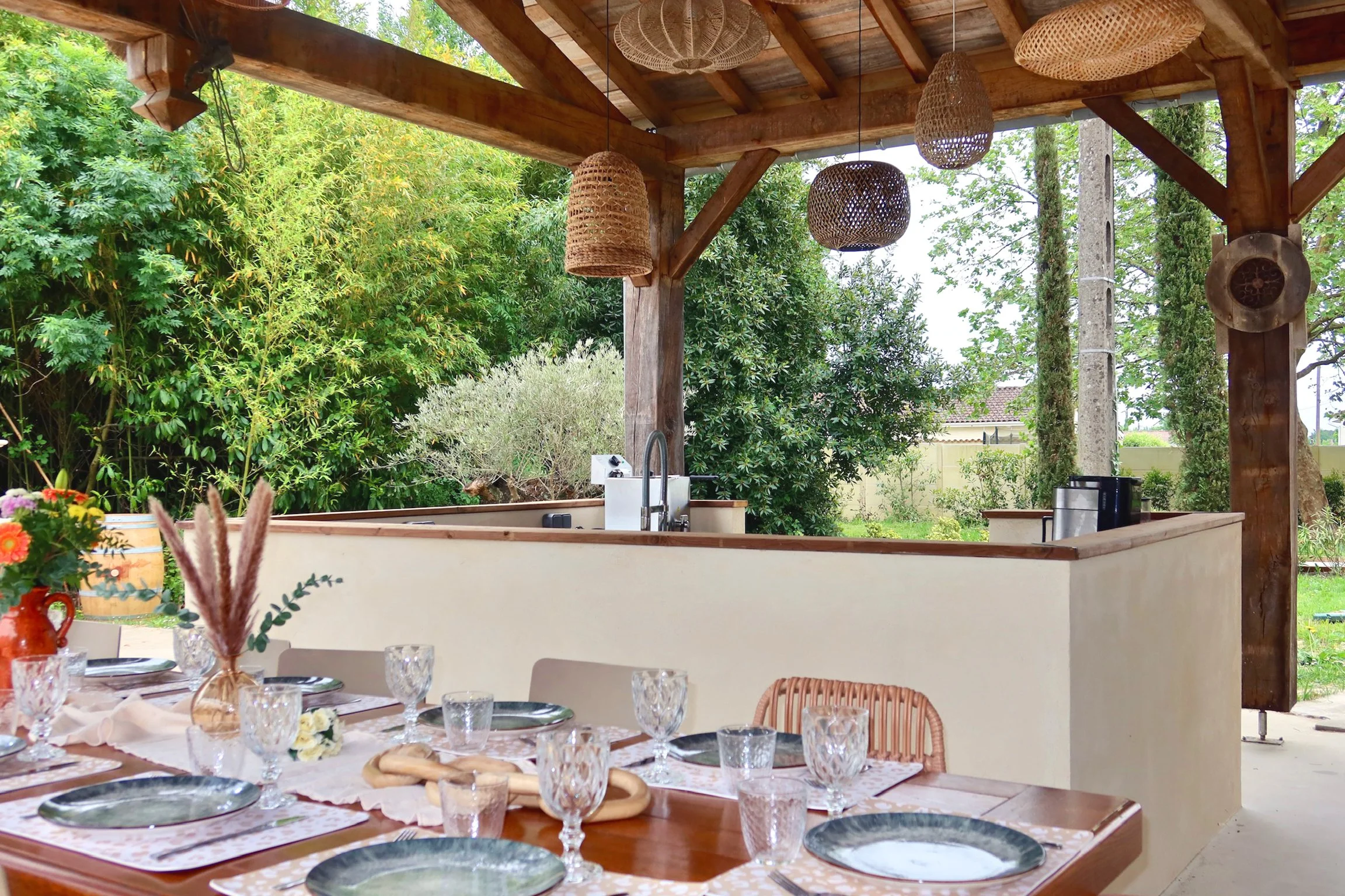 Une espace cuisine extérieure avec un plan de travail en bois, des décorations en rotin suspendues, et un jardin verdoyant en arrière-plan.