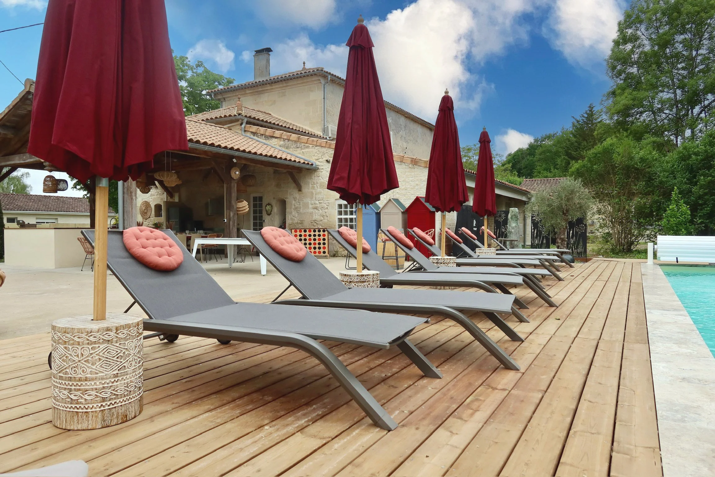 Chaises longues avec des coussins roses sous des parasols rouges près d'une piscine dans un espace extérieur avec un bâtiment en pierre en arrière-plan et des arbres verts autour.