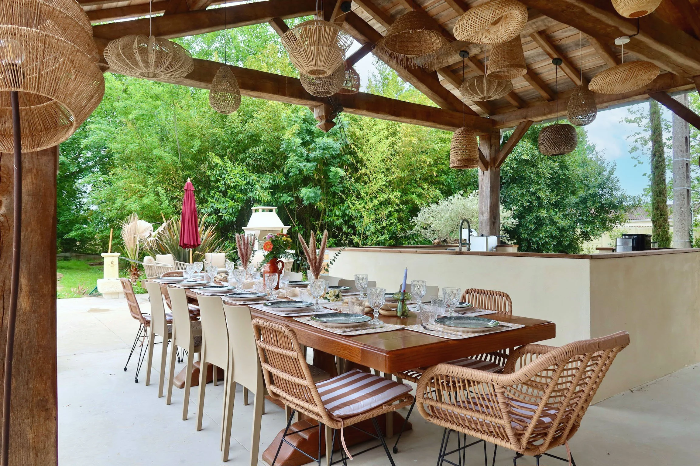 Une table de restaurant extérieure élégante, dressée avec des assiettes, des verres et des décorations florales, sous un toit en bois avec des luminaires en osier, entourée de verdure luxuriante.