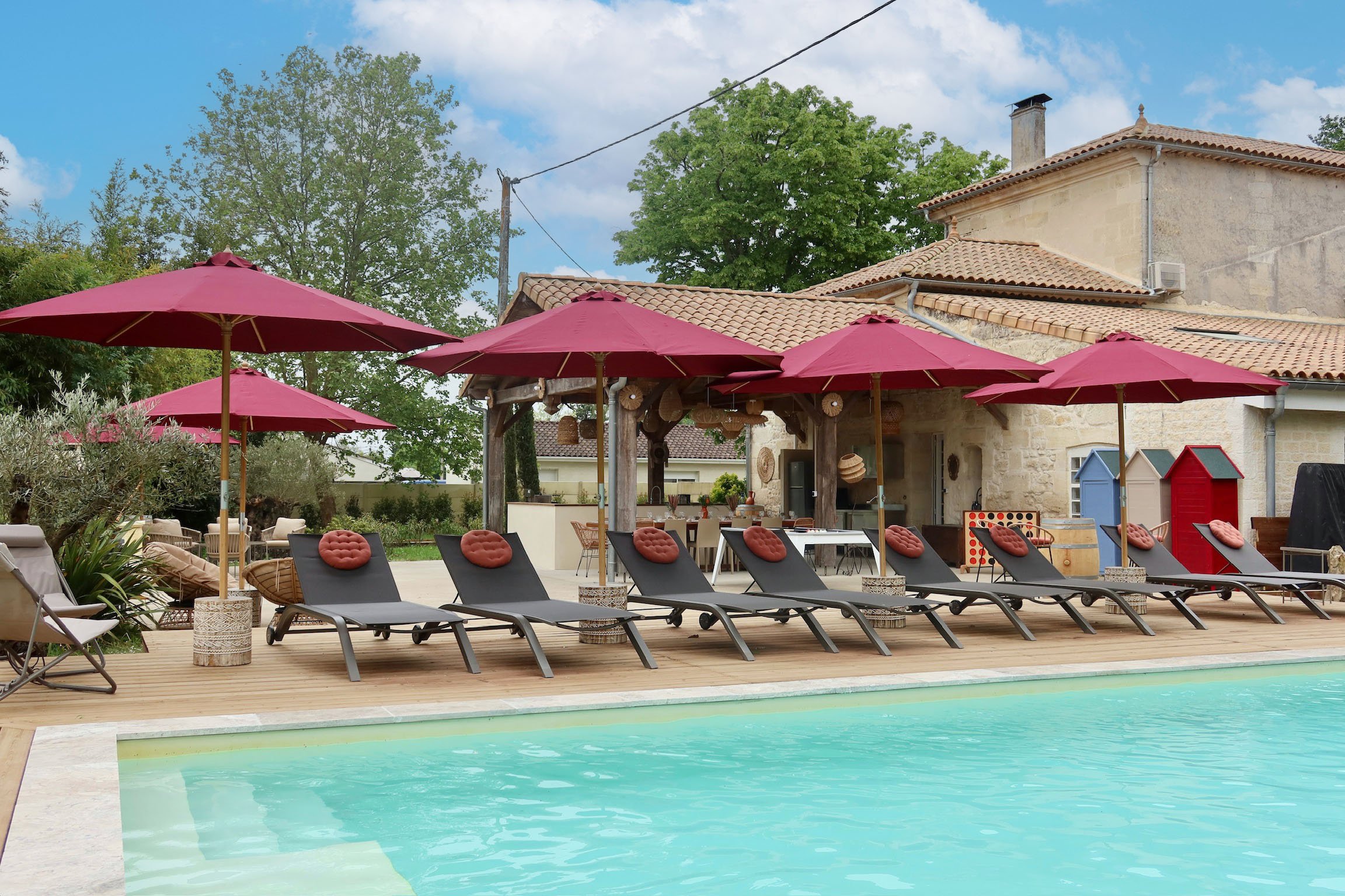 Une piscine extérieure avec des chaises longues équipées de coussins ronds, des parasols rouges, un espace lounge, et une maison en pierre en arrière-plan dans un jardin avec des arbres et un ciel partiellement nuageux.