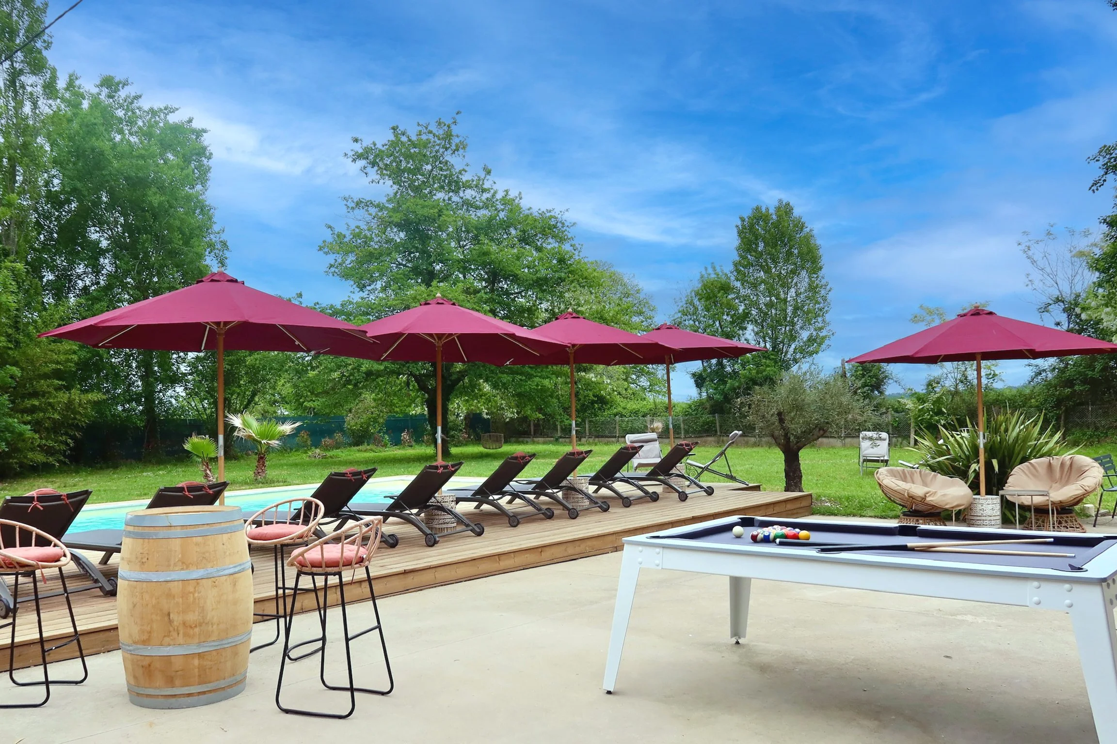 Un espace extérieur avec des parasols roses, des chaises longues, un billard pool, et un mobilier de jardin, entouré d'arbres verts sous un ciel bleu.