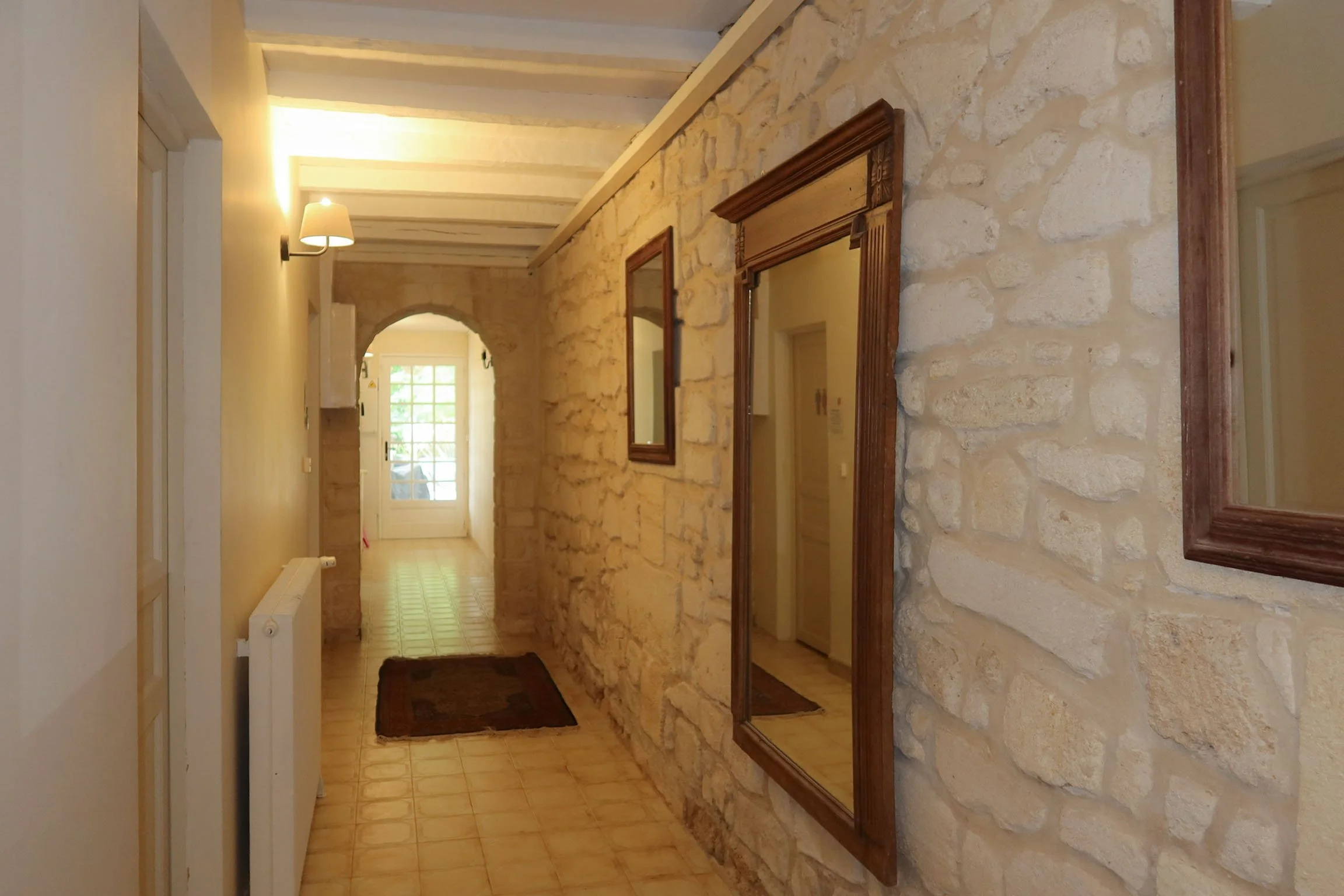 Intérieur d'une maison avec mur en pierre, miroir en bois, et porte fenêtre laissant entrer de la lumière naturelle.