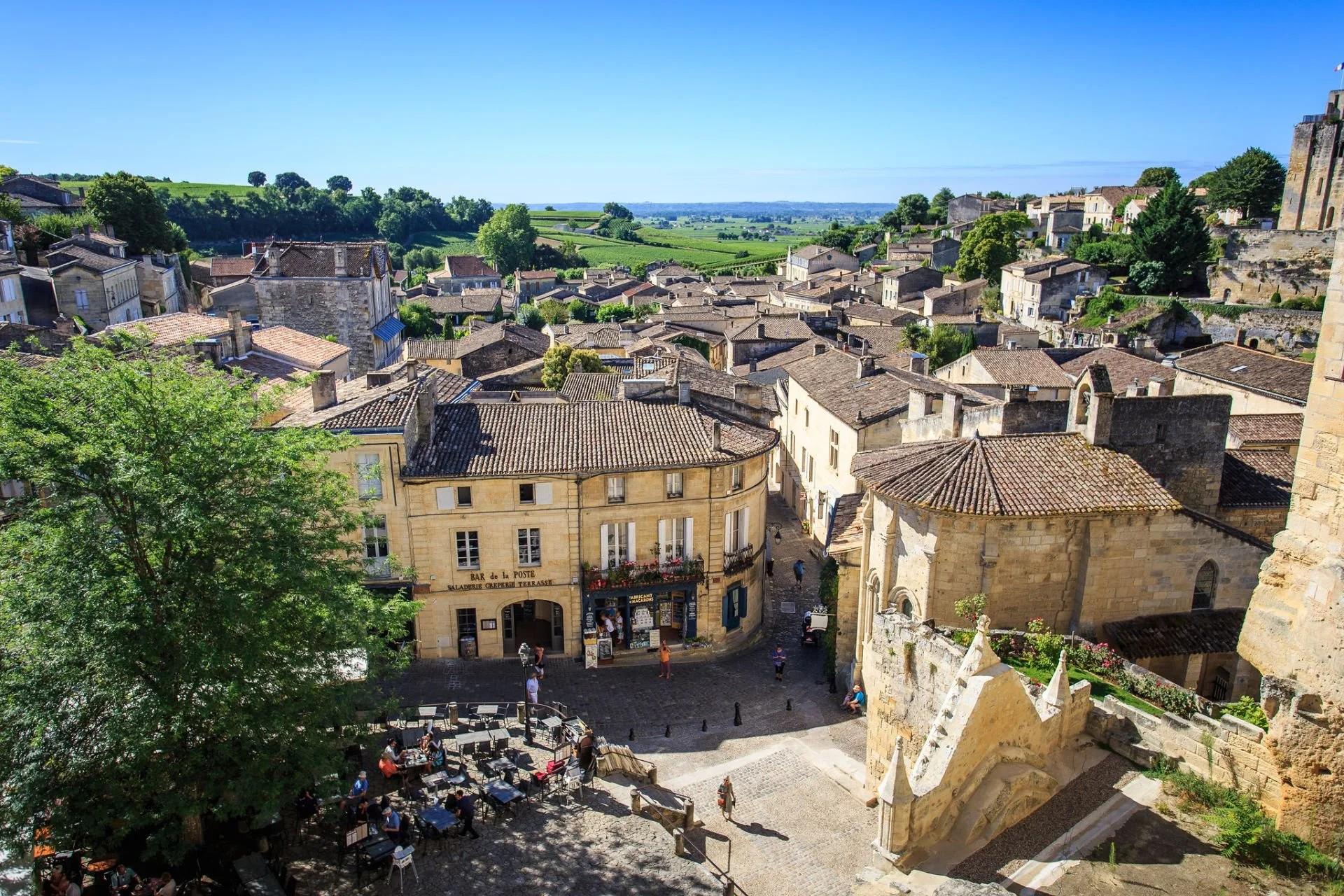 Vue d'une vieille ville méditerranéenne avec des toits en tuiles, des bâtiments en pierre, un café en plein air avec des tables, et un paysage verdoyant en arrière-plan.