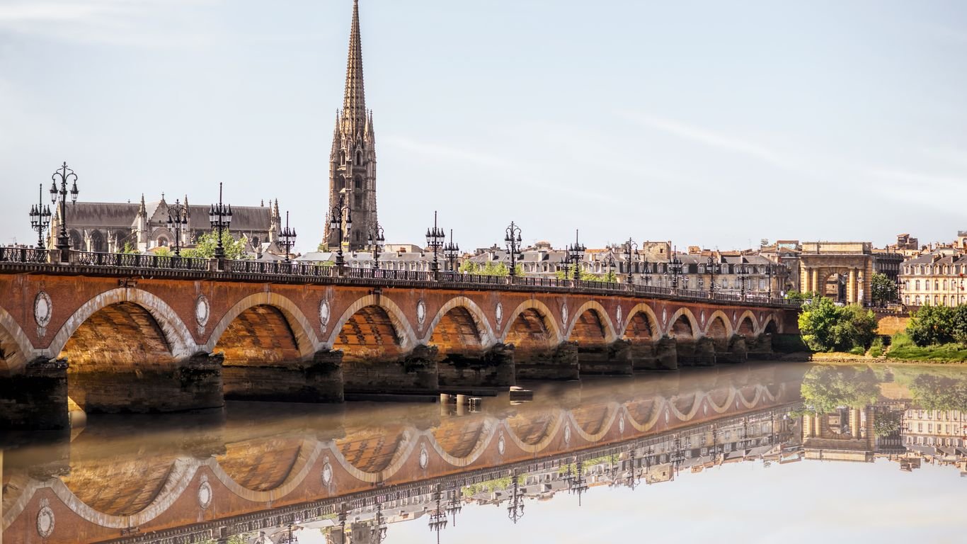Vue d'une ville avec un pont en pierre rouge sur une rivière, une église avec une haute flèche gothique, des lampadaires noirs et des bâtiments anciens comme le Pont Saint-Louis à Toulouse, France.