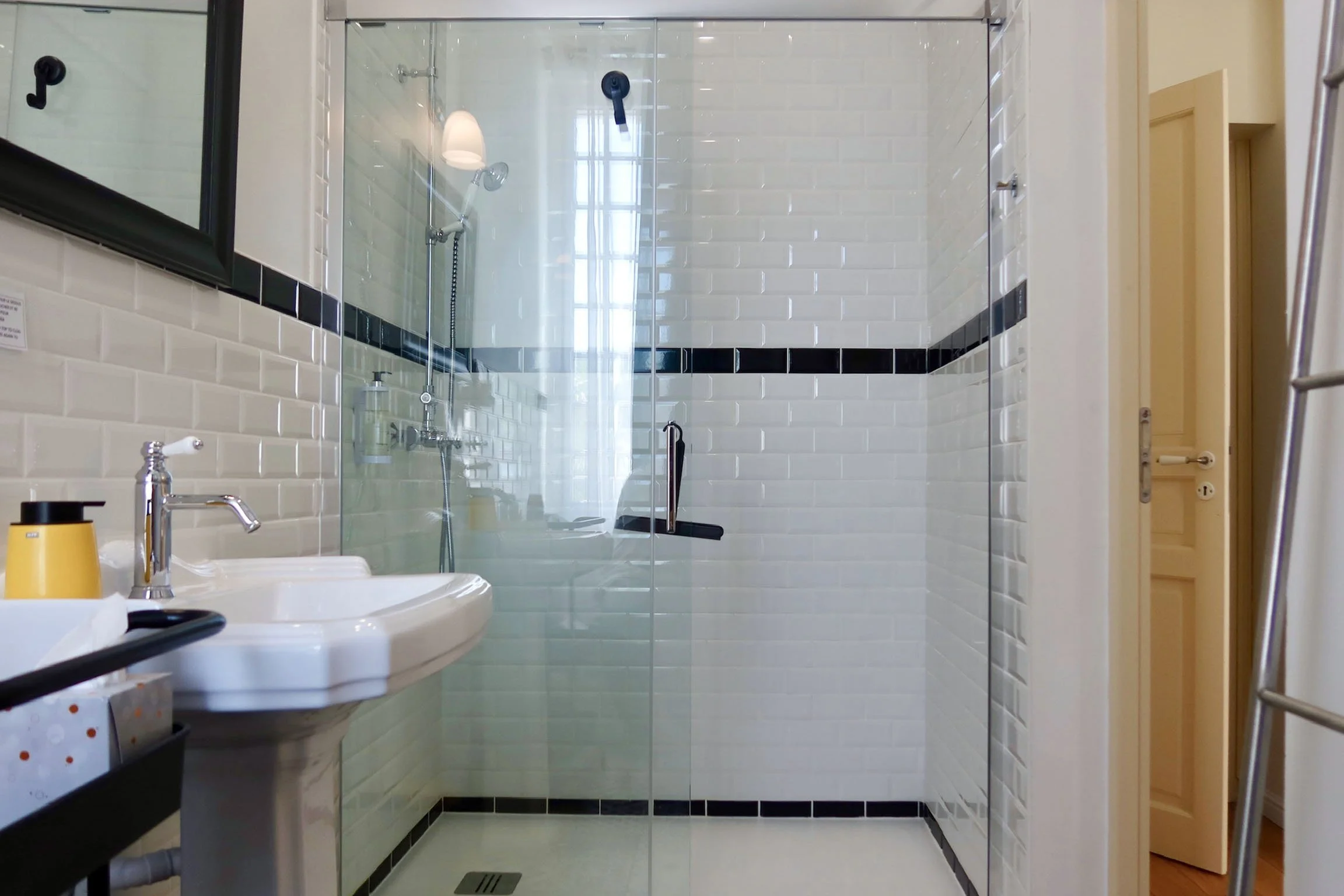 Salle de bain avec douche en verre, murs en carrelage blanc avec une bande de carrelage noir, lavabo avec distributeur de savon et miroir