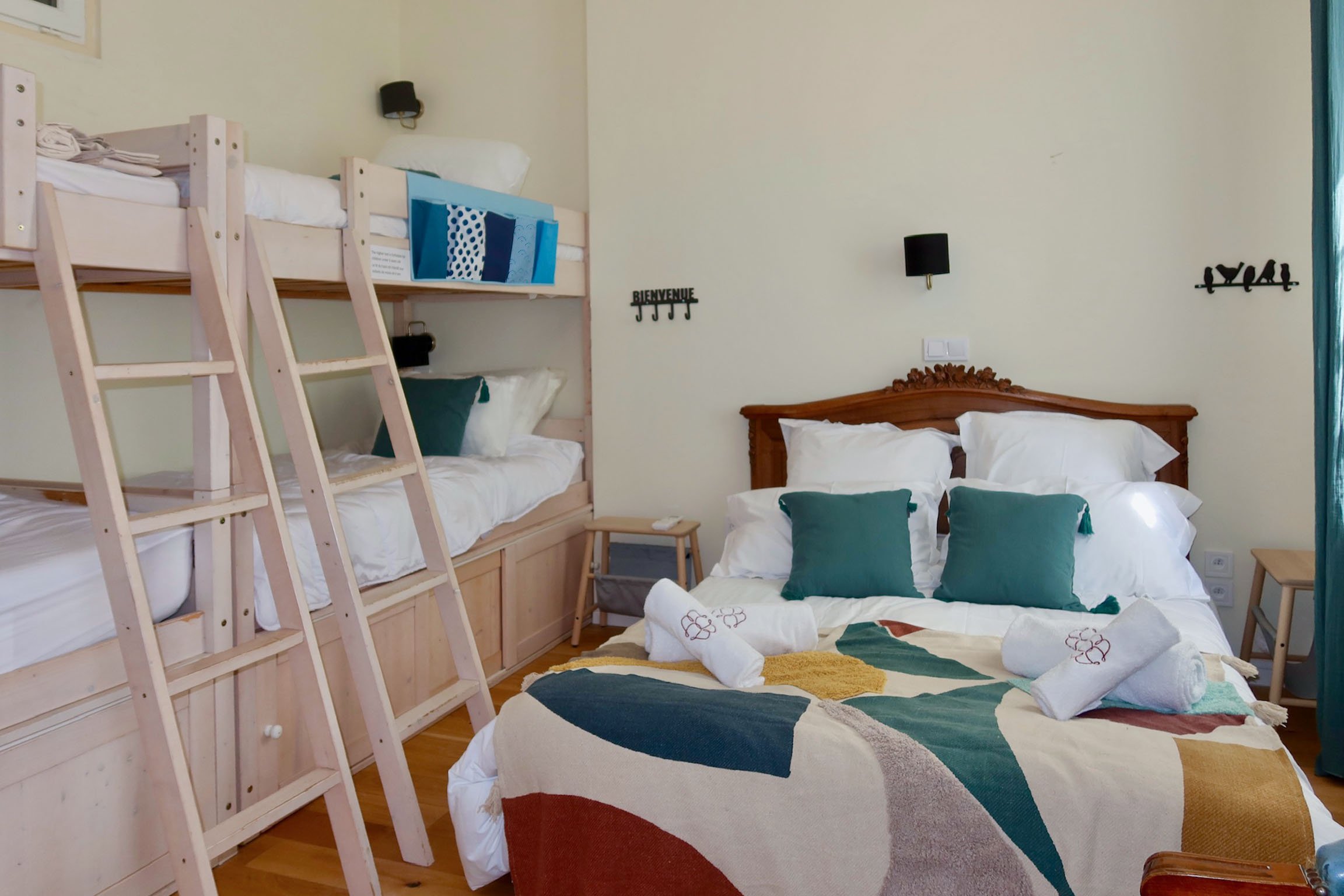 Chambre avec un grand lit, plusieurs oreillers, couvertures colorées, mur clair avec des décorations murales, et un lit mezzanine avec des couettes et coussins