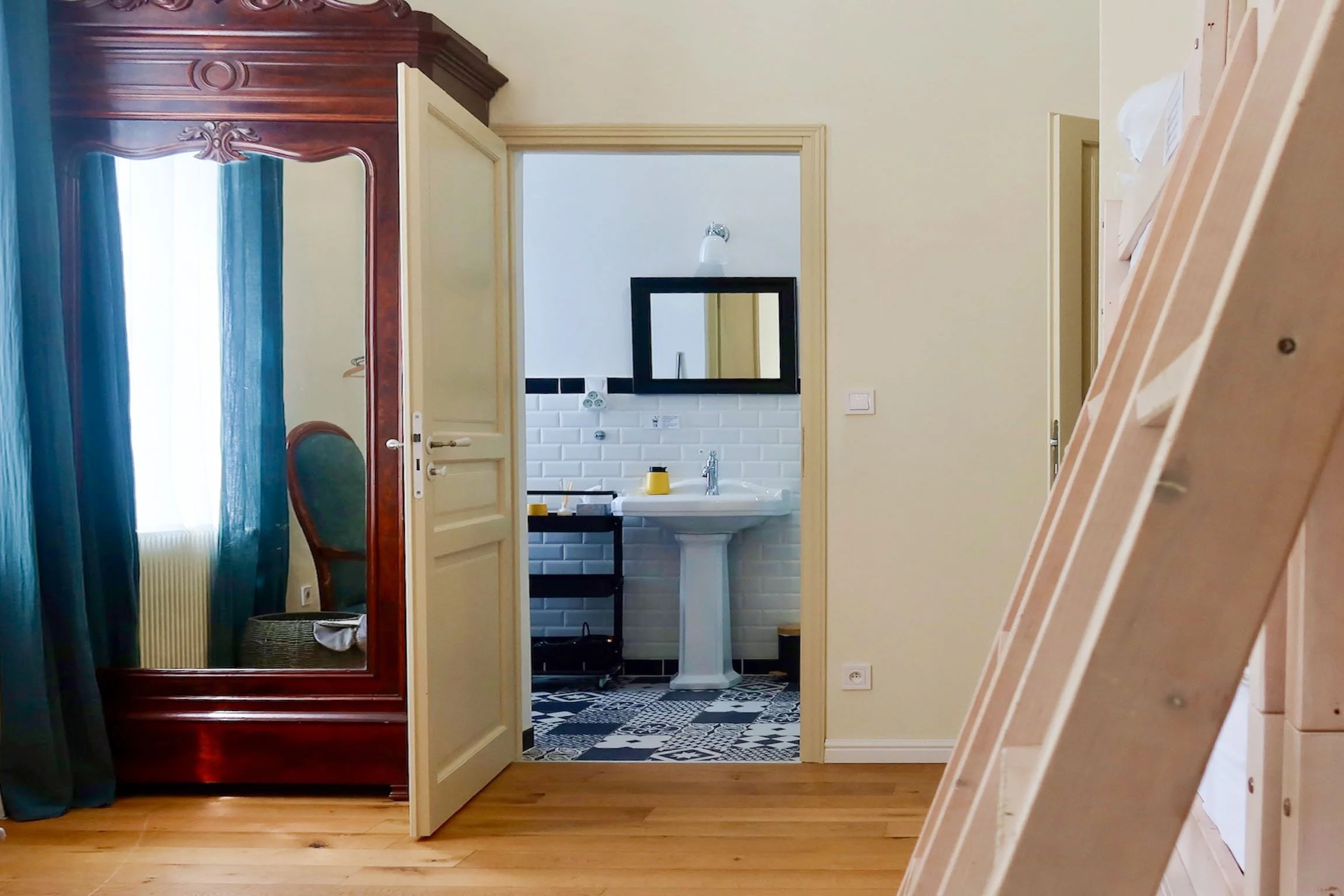 Vue d'un intérieur comprenant un miroir en bois, une porte ouverte menant à une salle de bain avec un lavabo, et un mur en brique blanche. Le sol est en bois clair.