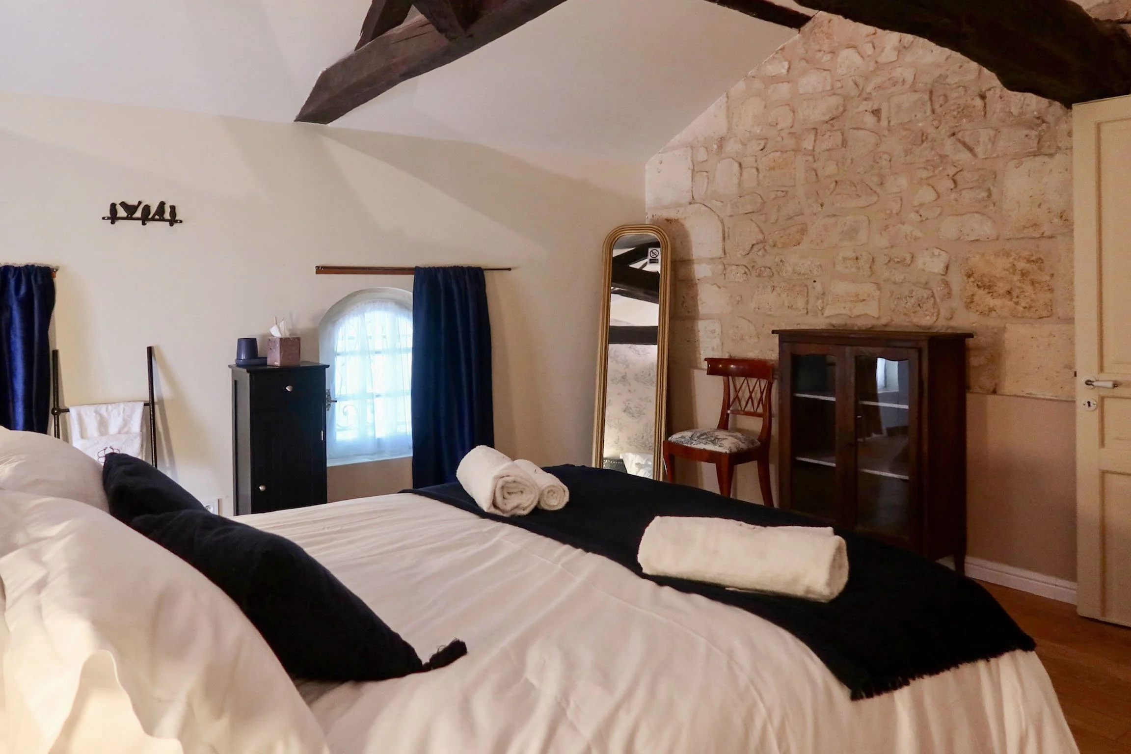 Chambre avec un lit double, deux serviettes blanches sur le lit, une fenêtre avec rideaux bleus, un miroir, un meuble en bois sombre, et un mur en pierre