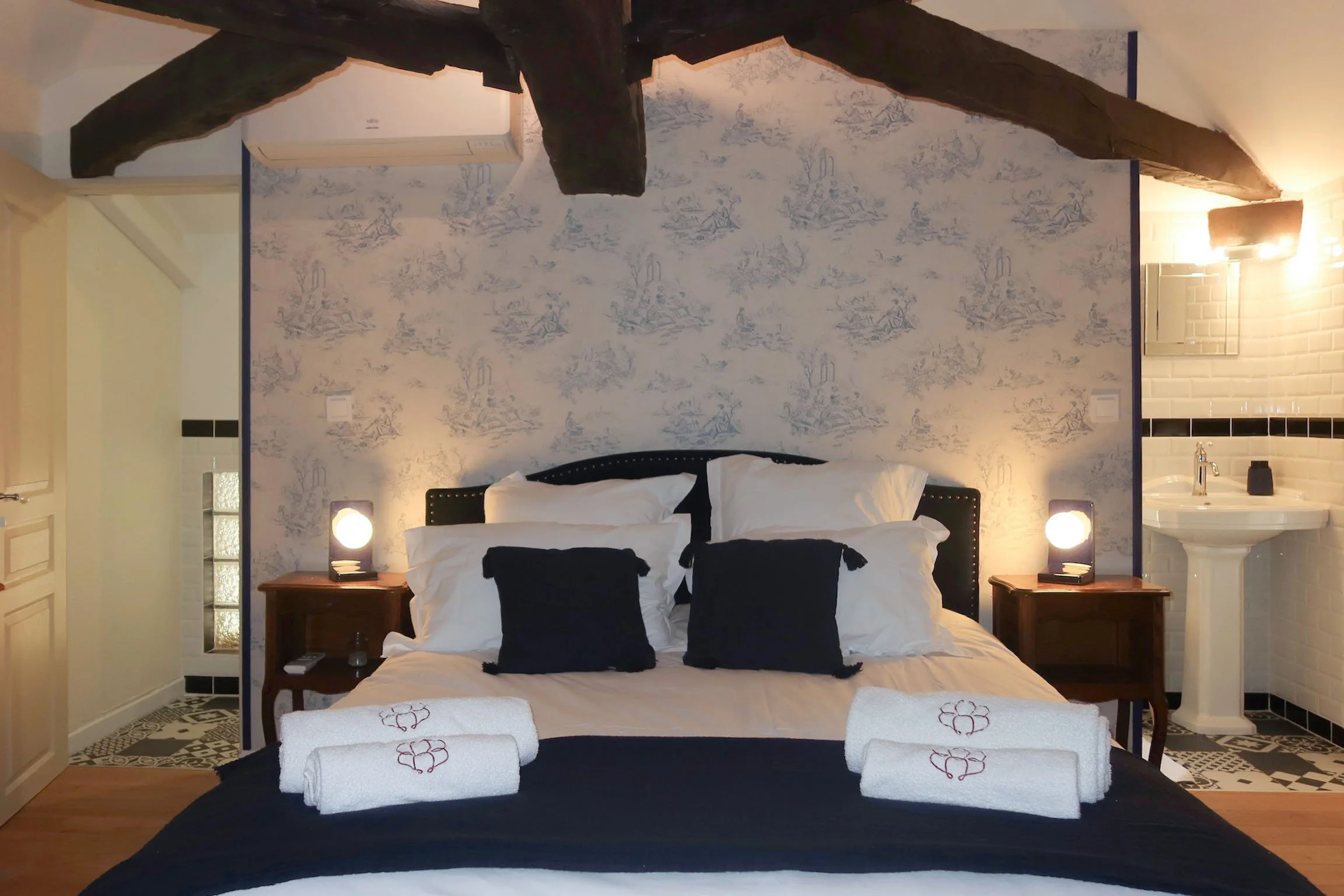 Lit double avec décoration noire et blanche, serviettes pliées sur le lit, lampes de lecture, mur avec papier peint à motifs bleus, petits meubles en bois, salle de bain partagée en arrière-plan.