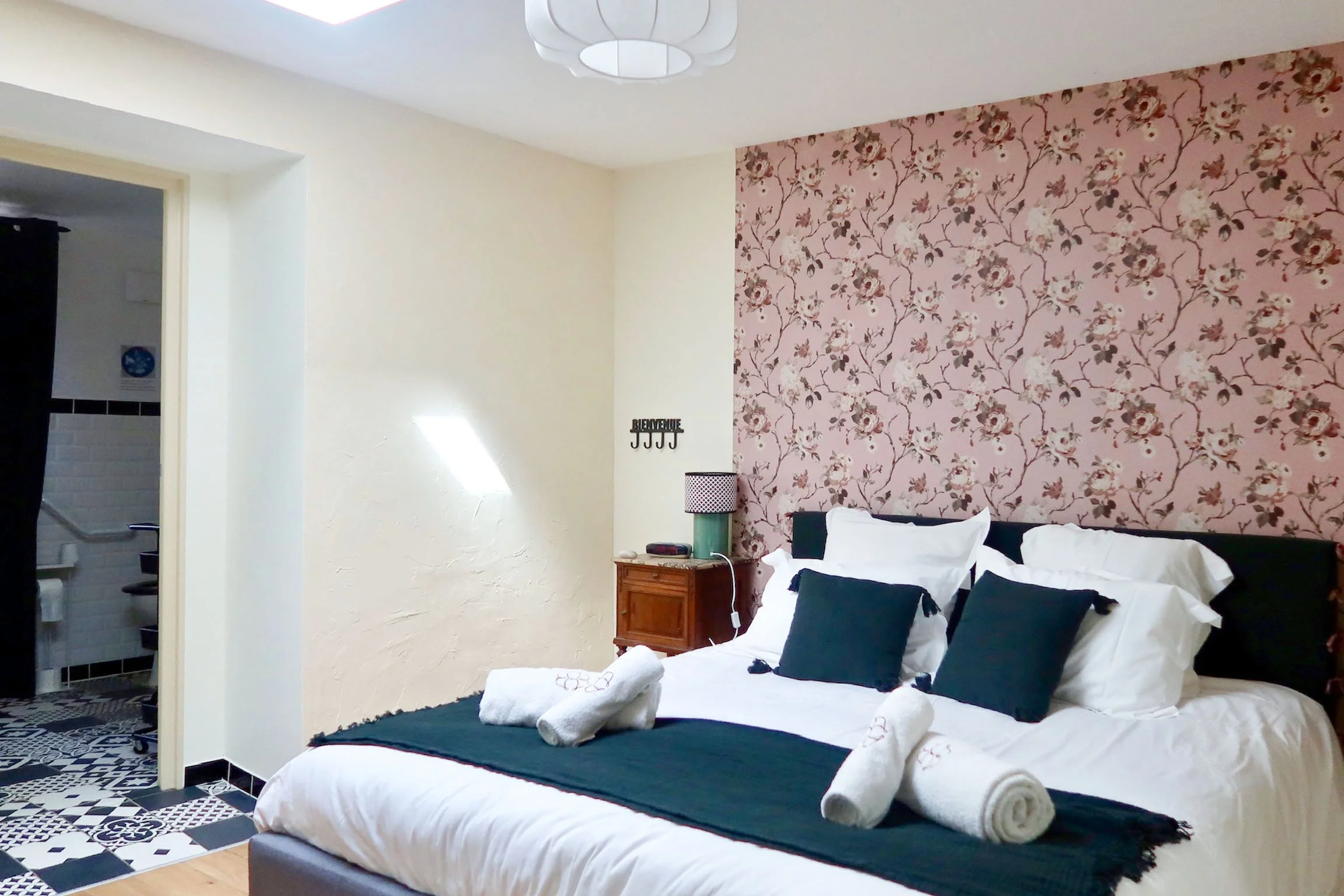 Chambre avec lit double, oreillers, serviettes enroulées, lampe de chevet, mur à motifs floraux roses, et porte menant à une salle de bain.