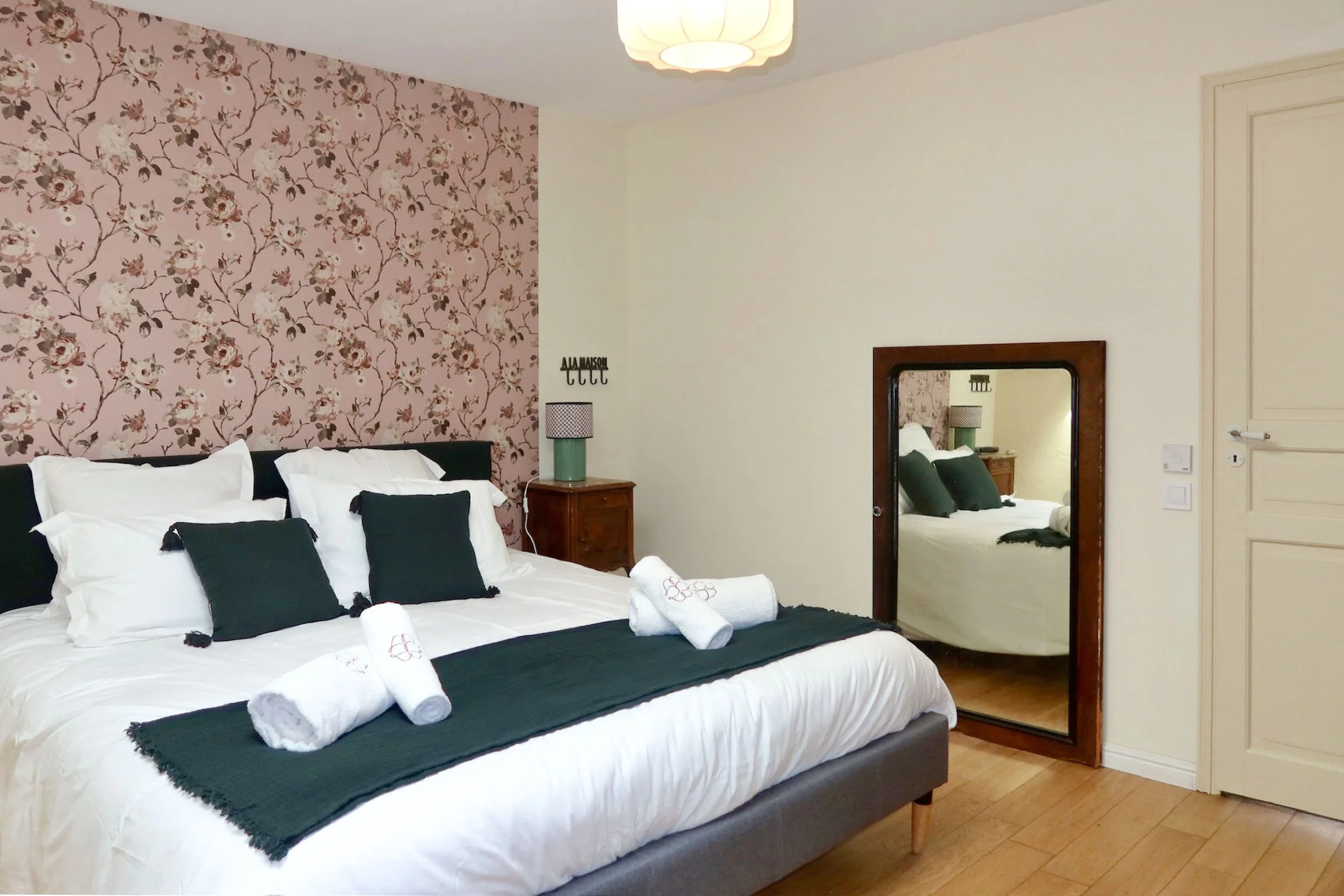 Chambre avec un lit double, draps blancs, coussins noirs, serviettes roulées sur le lit, lampe de chevet, mur à papier peint floral rose et beige, miroir en bois, porte et placard en bois, sol en parquet.