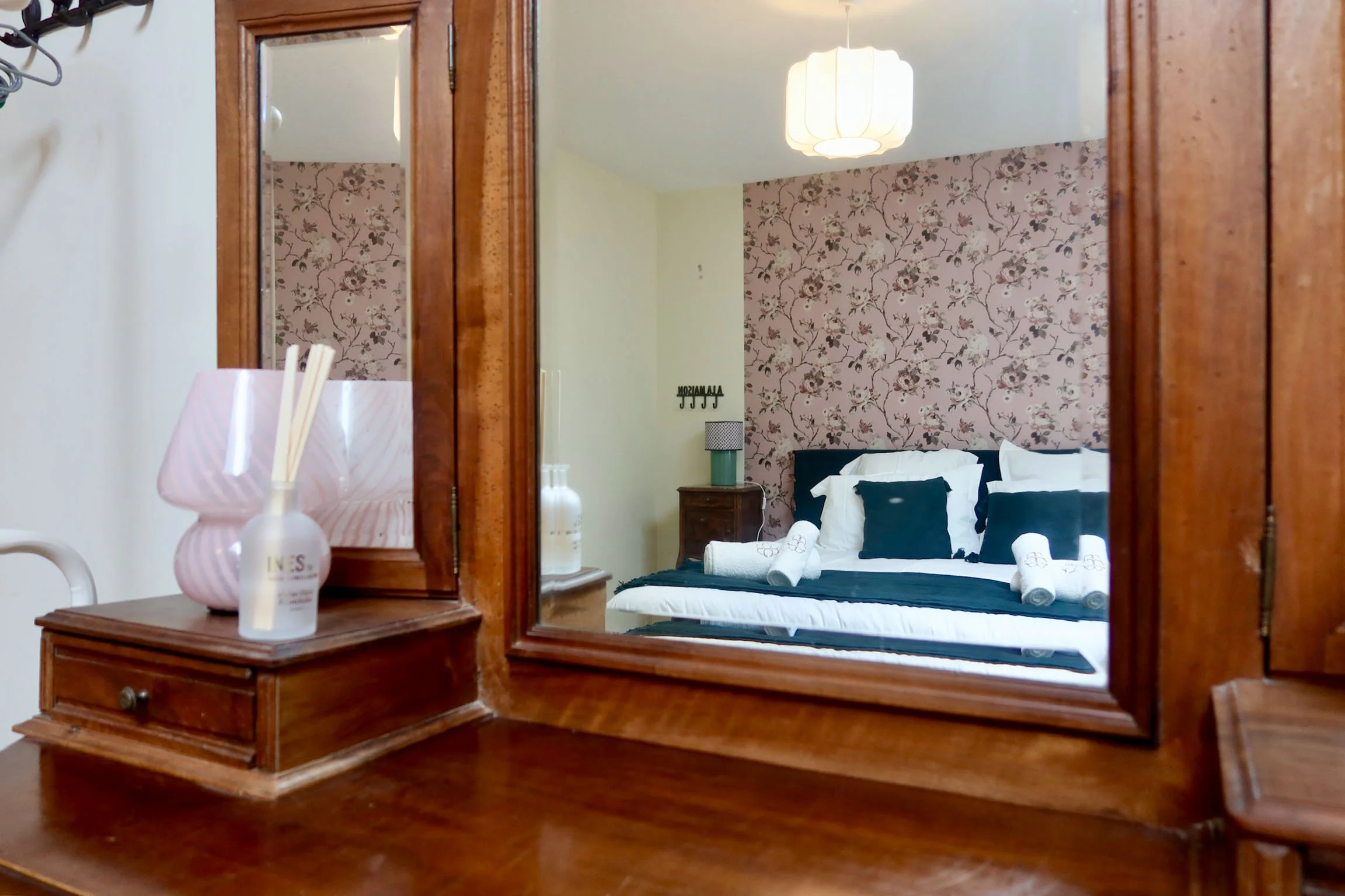 Un grand miroir en bois reflète une chambre à coucher avec un lit, des oreillers noirs et blancs, une lampe de chevet, un mur à papier peint floral et une lampe suspendue