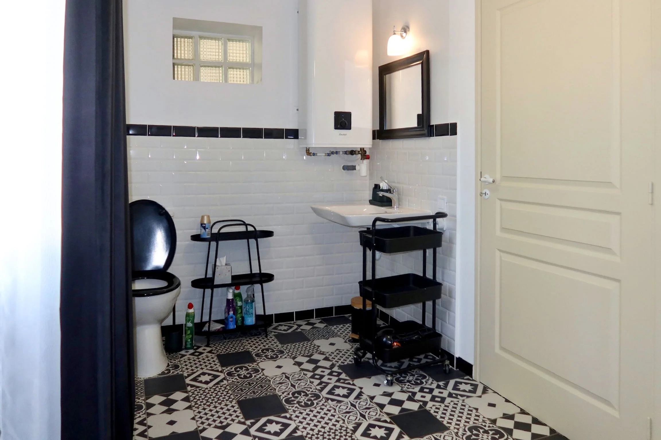 Une salle de bain avec un sol à carreaux noirs et blancs, un miroir noir au mur, un lavabo, un chauffage d'eau, et divers produits de nettoyage au sol.