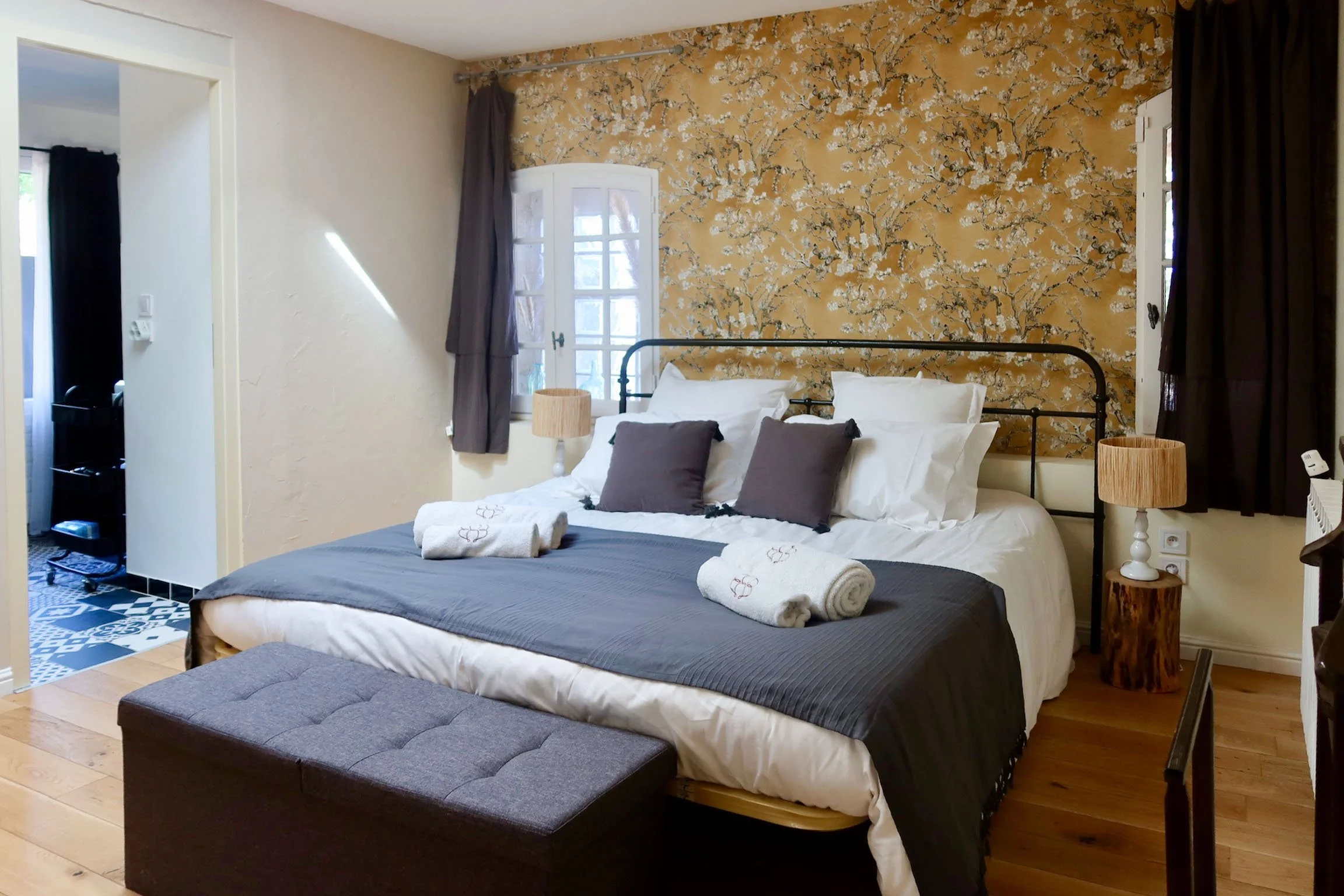 Chambre avec lit double, draps blancs et coussins gris, imprimé floral sur le mur derrière le lit, deux lampes de chevet en bois, un banc gris au pied du lit, fenêtres avec rideaux gris, sol en bois, coin toilettes visible à gauche.