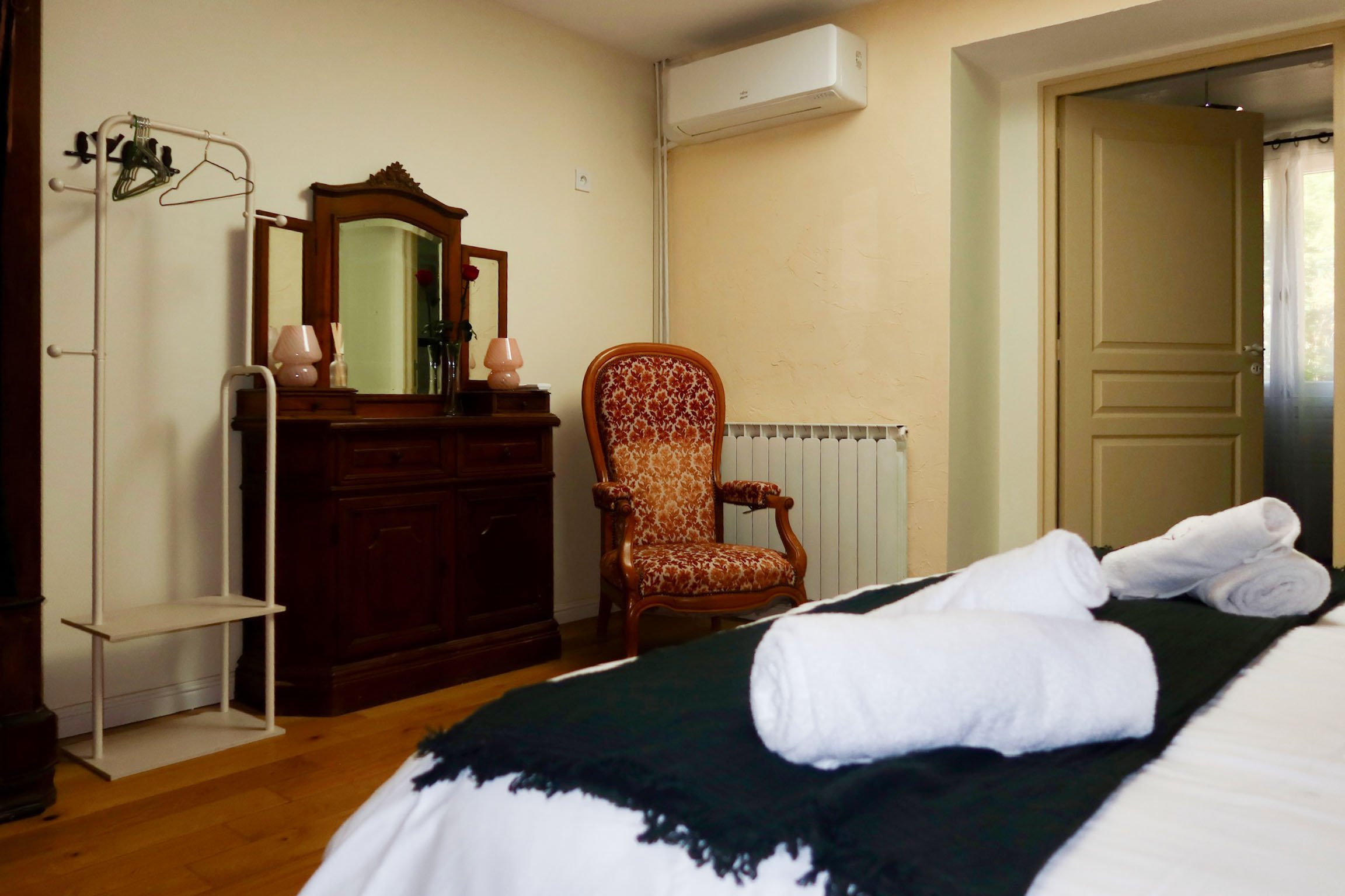 Chambre avec un lit, un miroir ancien, une chaise en bois avec tissu rouge à motifs, une lampe rose, un miroir mural, un climatiseur, une porte entrouverte et une fenêtre avec un rideau blanc.