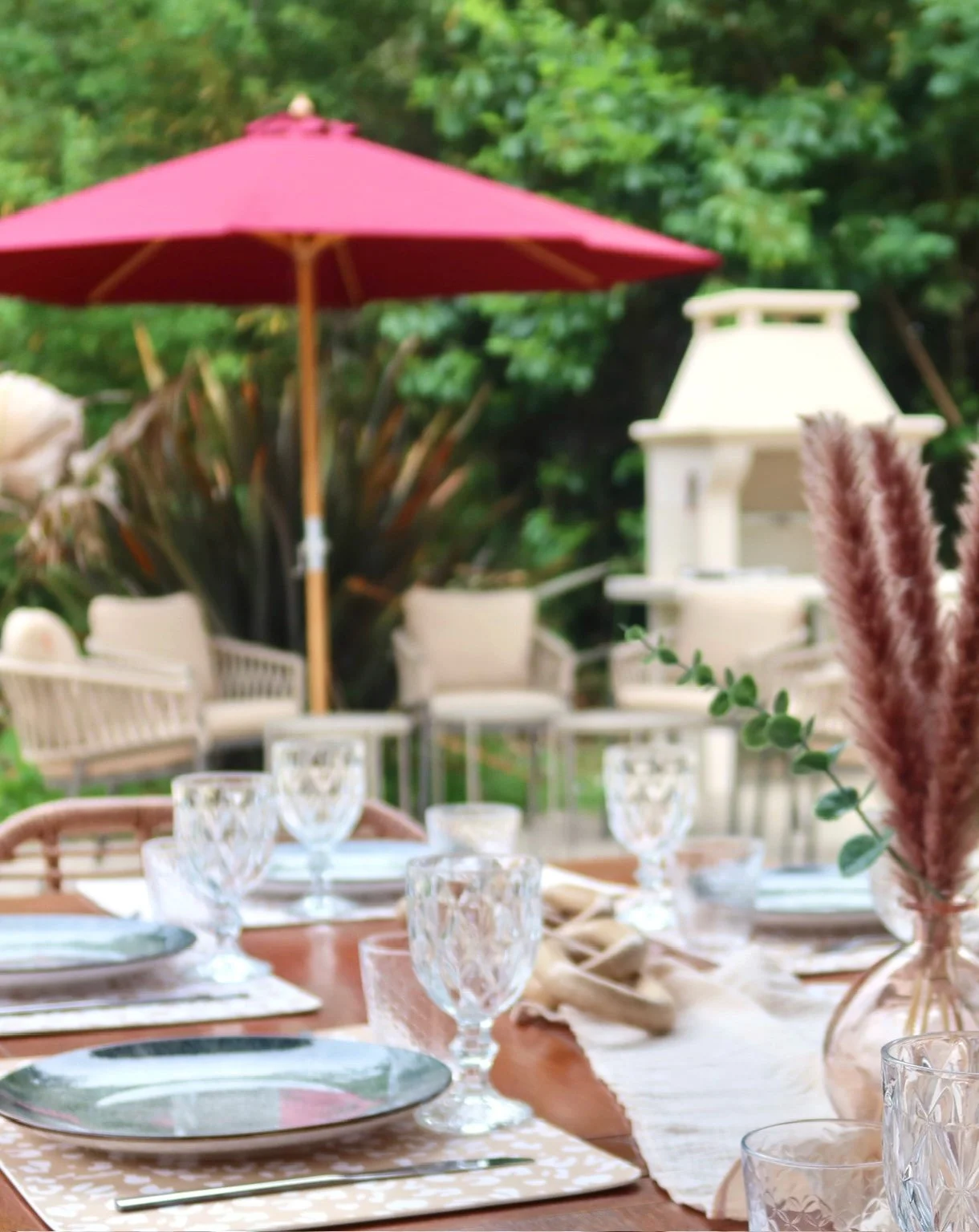 Table dressée pour un repas en plein air avec des verres à vin, assiettes, et un centre de table avec des pampas. En arrière-plan, un parasol rouge, un barbecue blanc, et un jardin verdoyant.