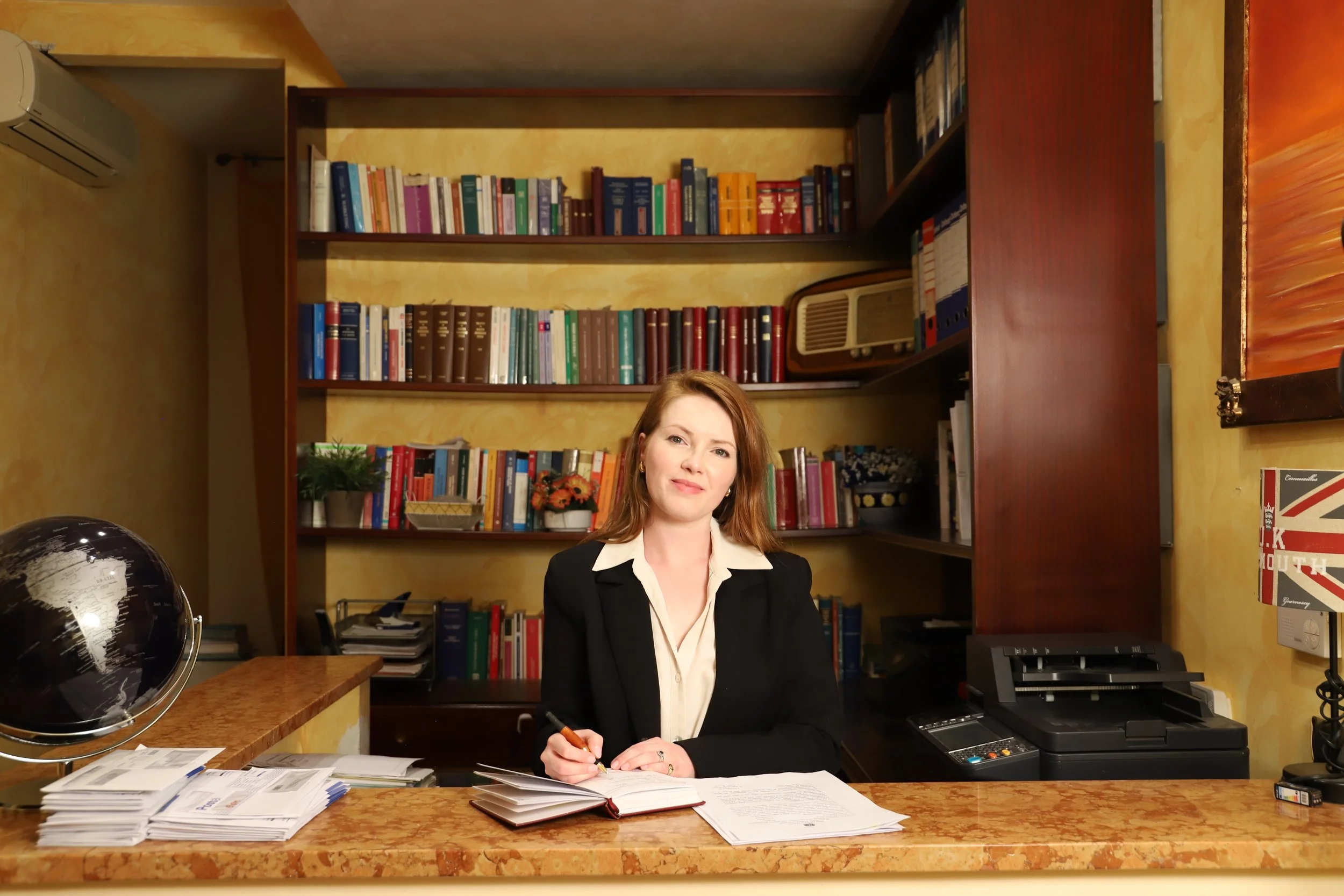 A woman sitting at a desk in an office, surrounded by bookshelves filled with books. She is wearing a black blazer and white shirt, holding a pen, and appears to be working or preparing for a meeting.