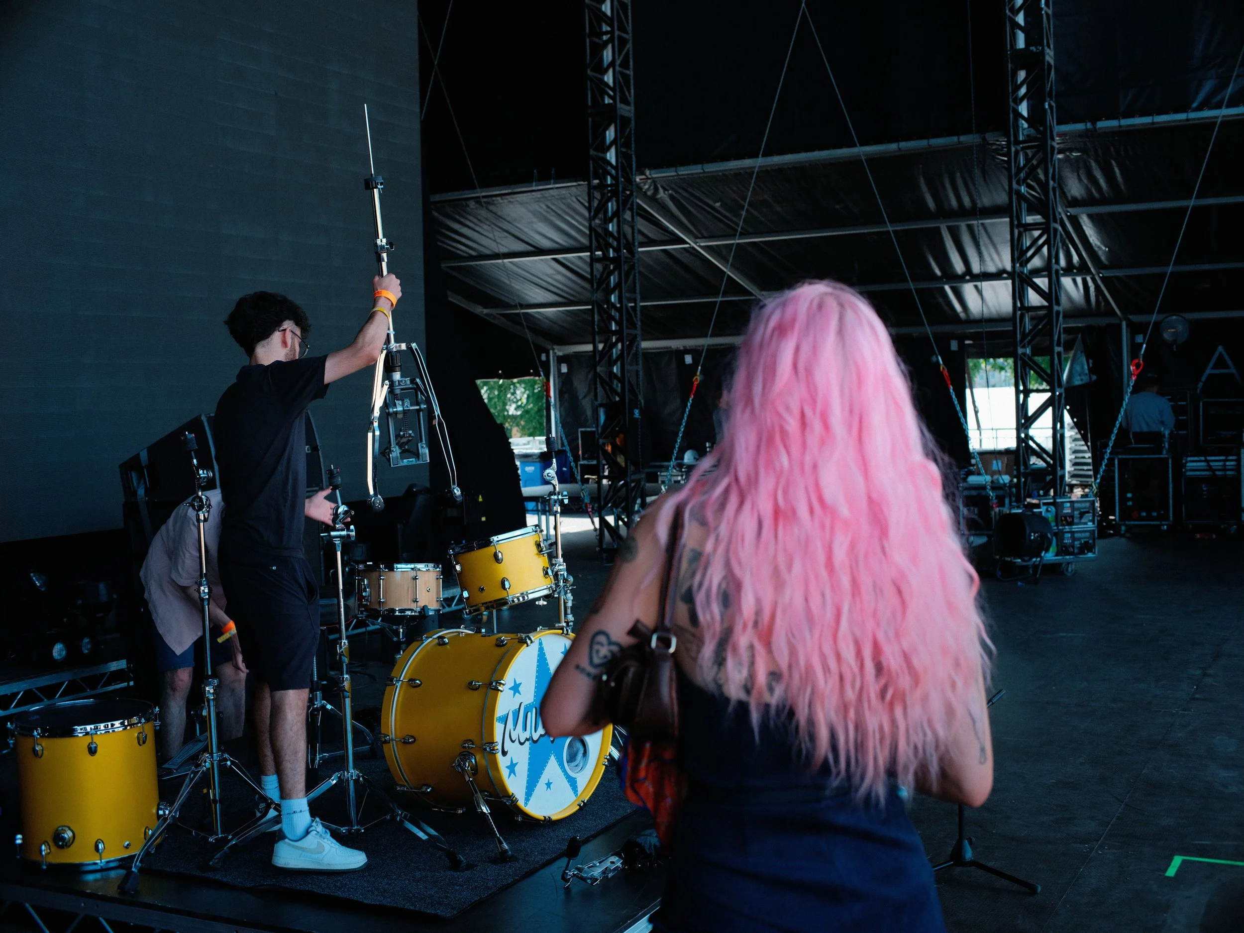 A person with pink hair sitting in front of a stage with a drummer setting up drum equipment, including a yellow bass drum with a star design, in an outdoor concert venue.