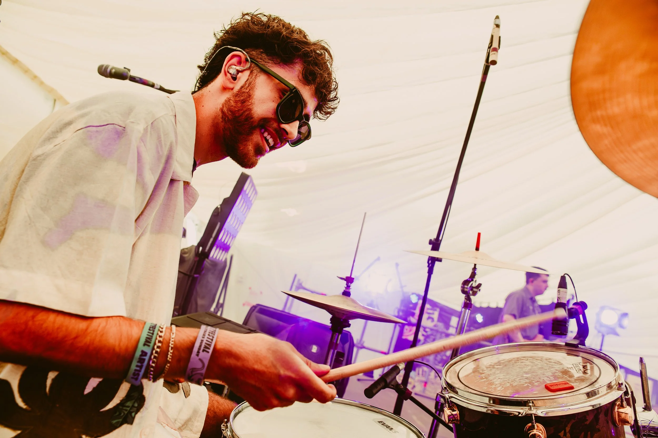 A man with a beard, wearing sunglasses and bracelets, playing drums at an indoor event with a white tent ceiling and electronic equipment in the background.