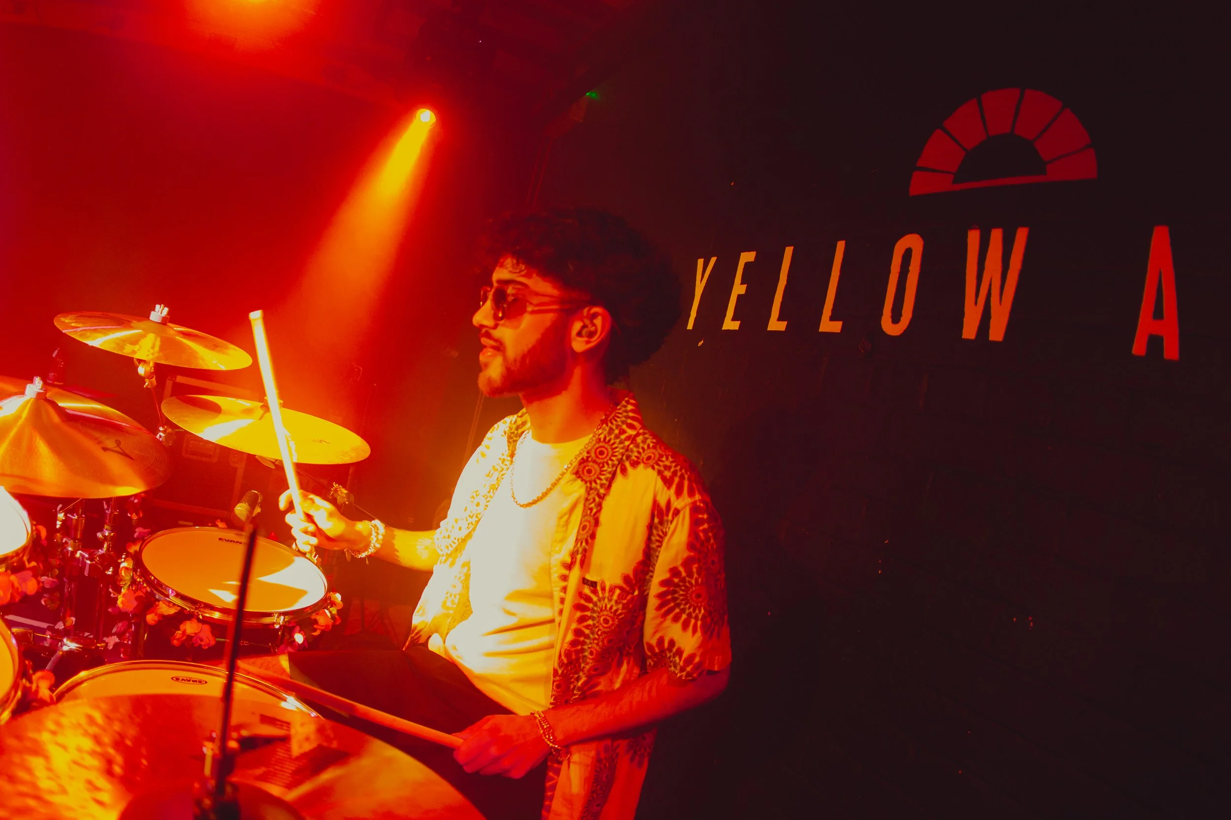 A man playing drums on stage illuminated by red and yellow stage lights, with a dark background featuring the words 'YELLOW A' and a semi-circle logo.